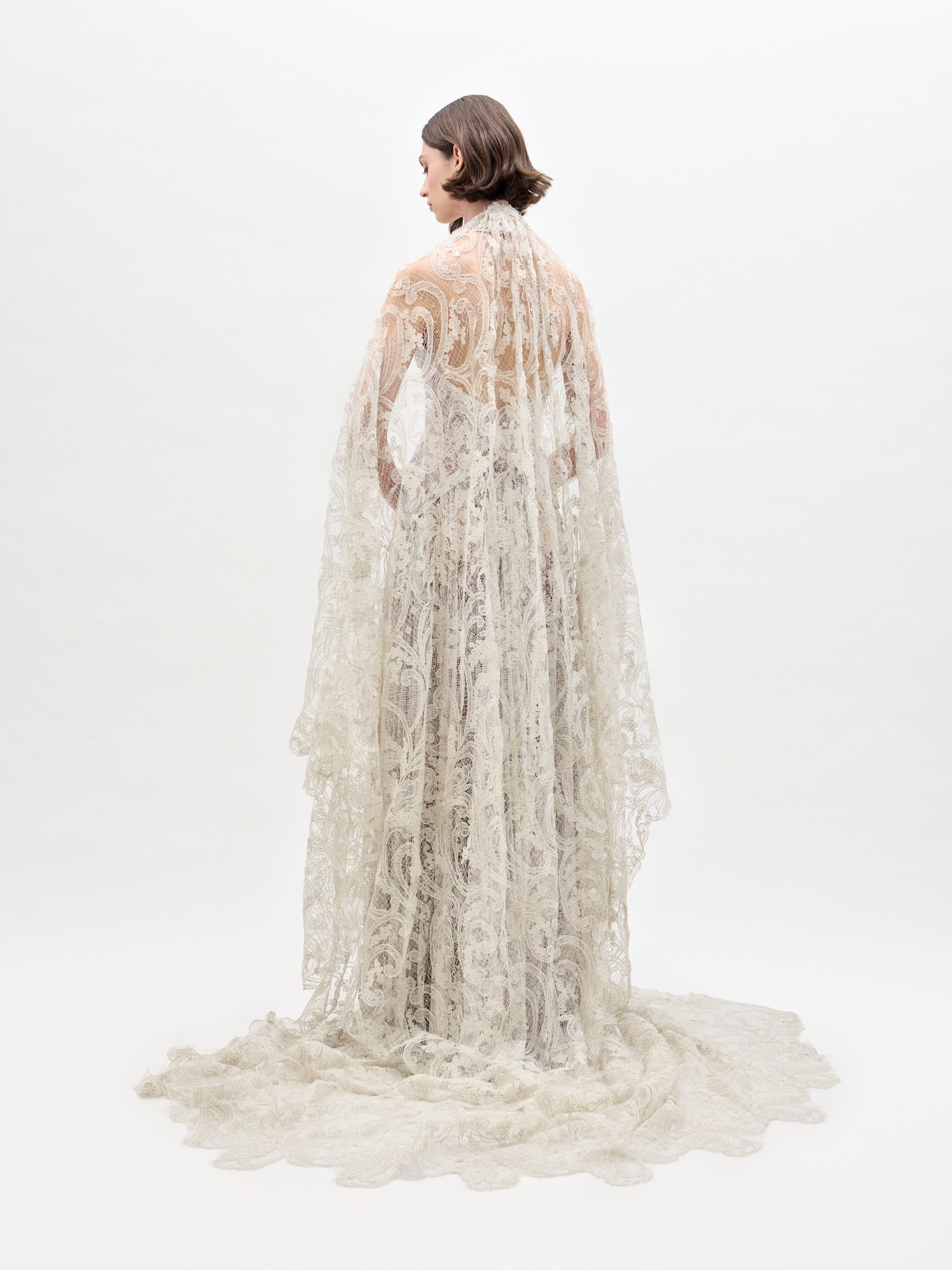 Wearing the Ilana Dress, a woman in a long lace bridal gown holds a bouquet of white and purple flowers against a plain background.