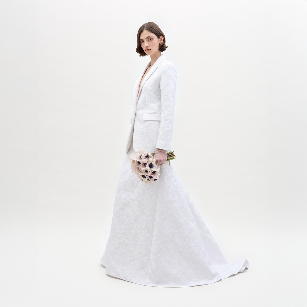 Wearing an Imola Skirt, a person in a white textured suit holds a bouquet of light-colored flowers against a plain background.