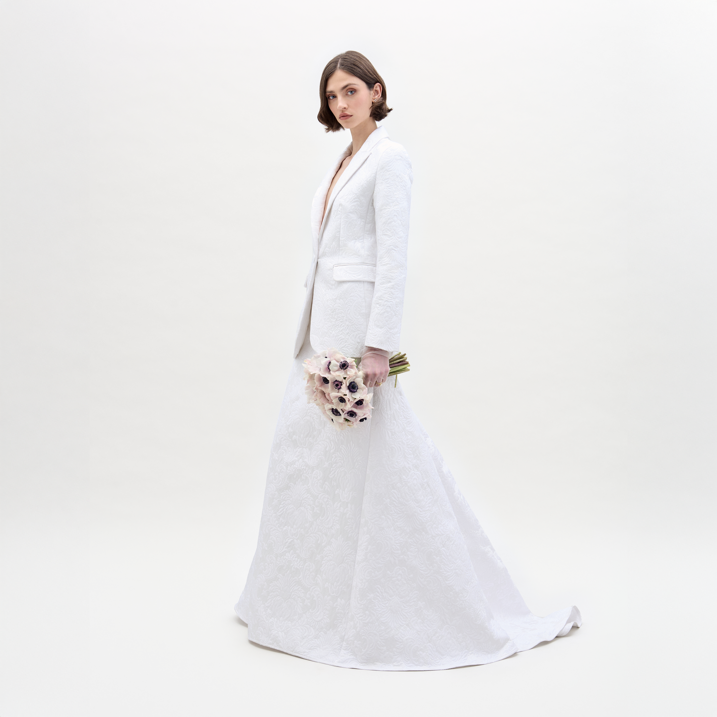 Wearing an Imola Skirt, a person in a white textured suit holds a bouquet of light-colored flowers against a plain background.