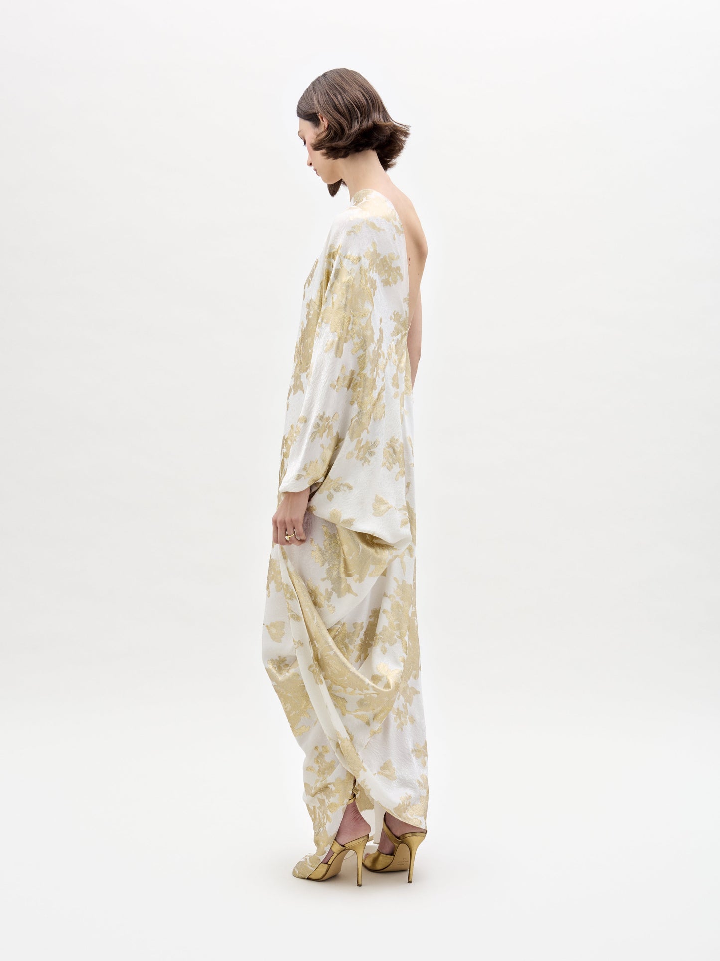 A woman dons the elegant Inu Dress, a flowing white and gold attire with a floral pattern, standing against a plain white background.