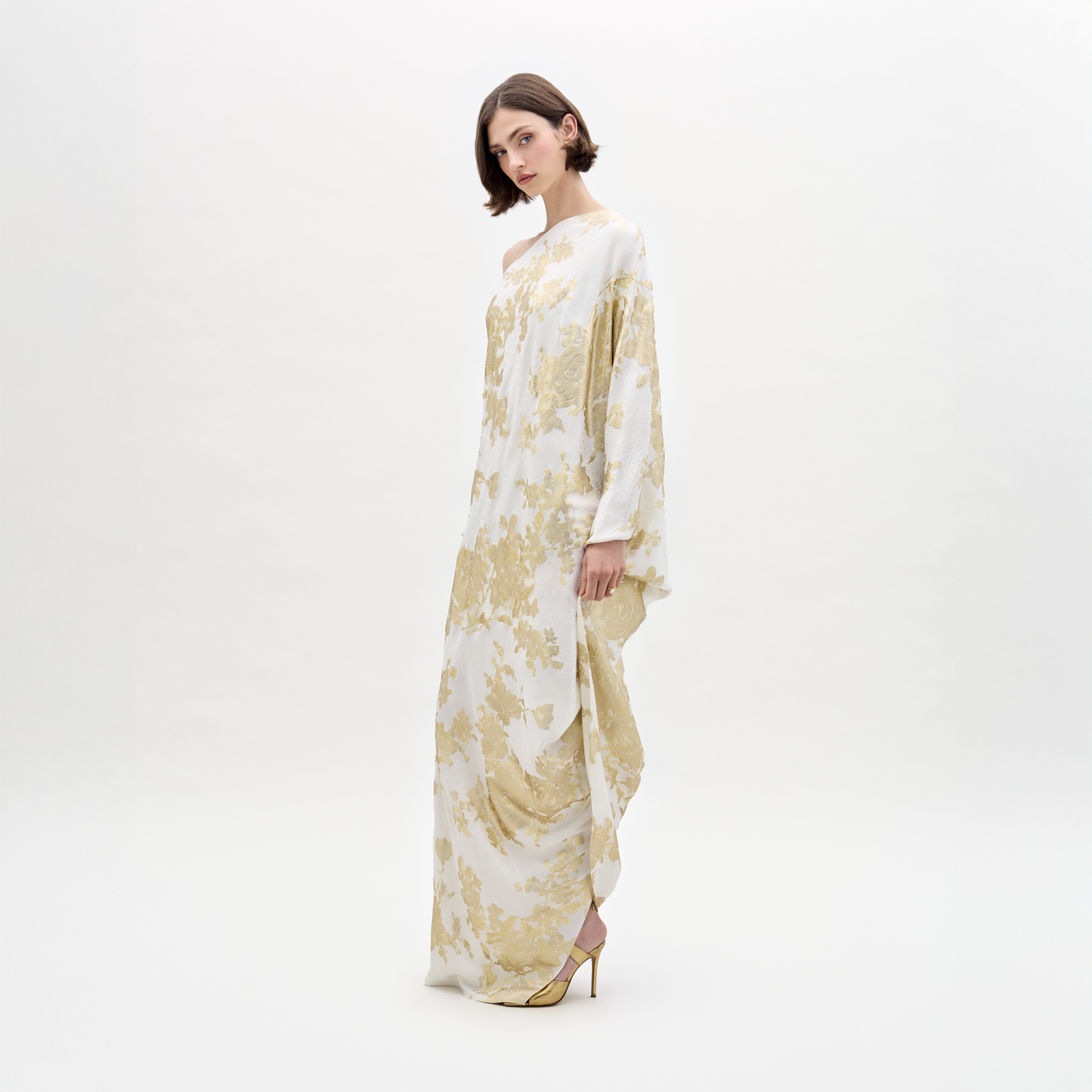 A woman dons the elegant Inu Dress, a flowing white and gold attire with a floral pattern, standing against a plain white background.