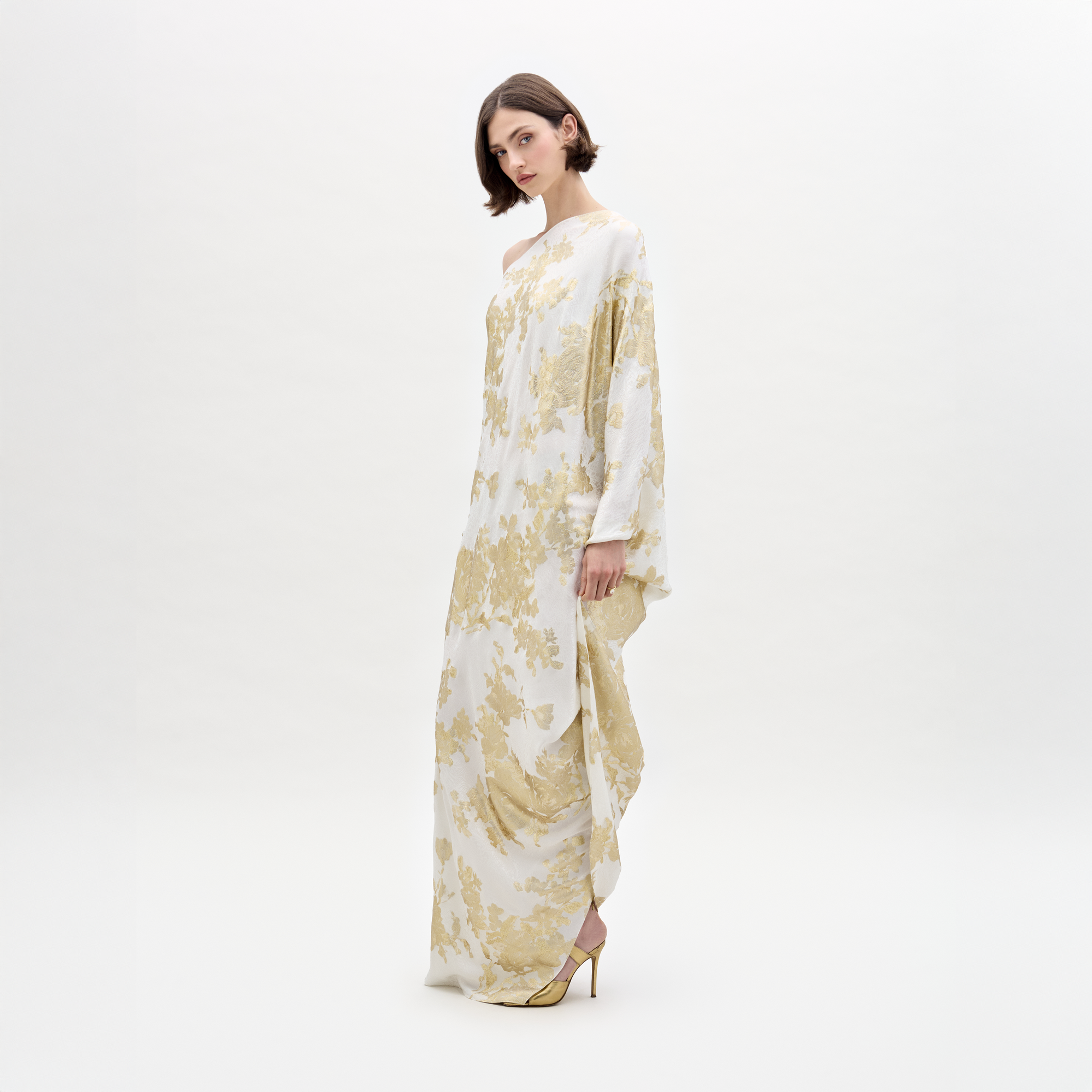 A woman dons the elegant Inu Dress, a flowing white and gold attire with a floral pattern, standing against a plain white background.