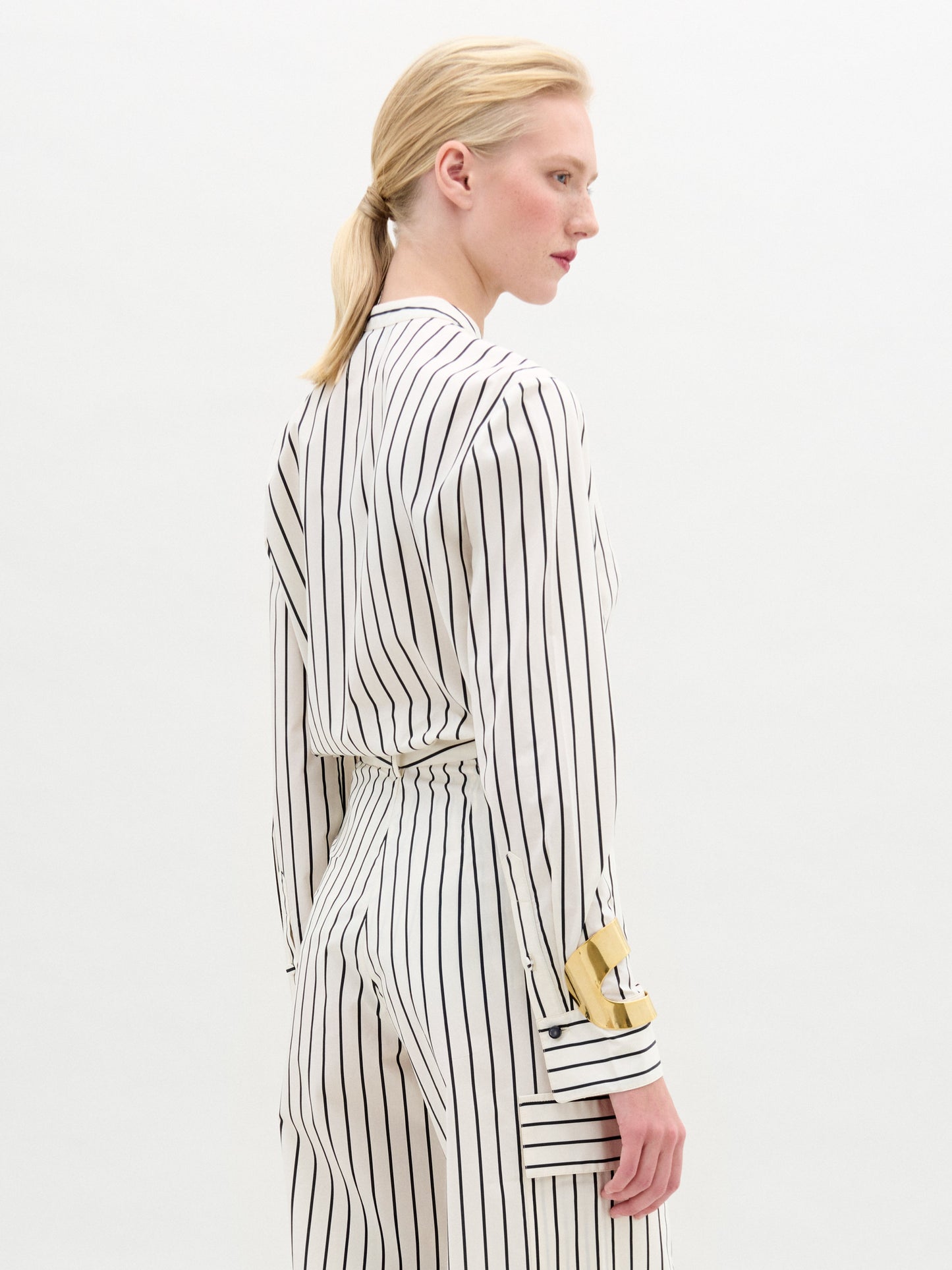 A person stands against a plain background wearing the Isabell Blouse, a white long-sleeve top with black lace and delicate embroidery down the front, paired with pants featuring black vertical stripes.