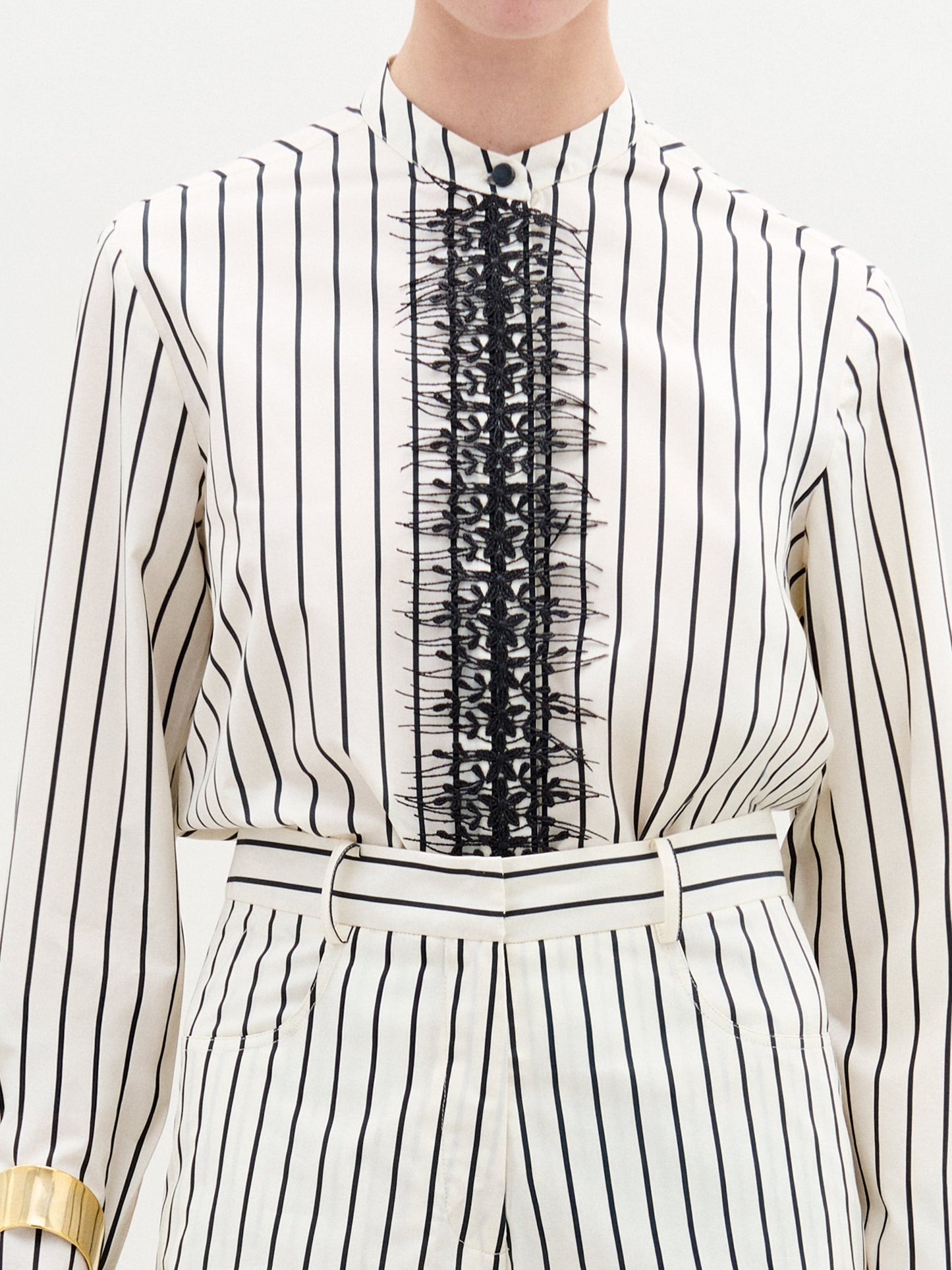 A person stands against a plain background wearing the Isabell Blouse, a white long-sleeve top with black lace and delicate embroidery down the front, paired with pants featuring black vertical stripes.