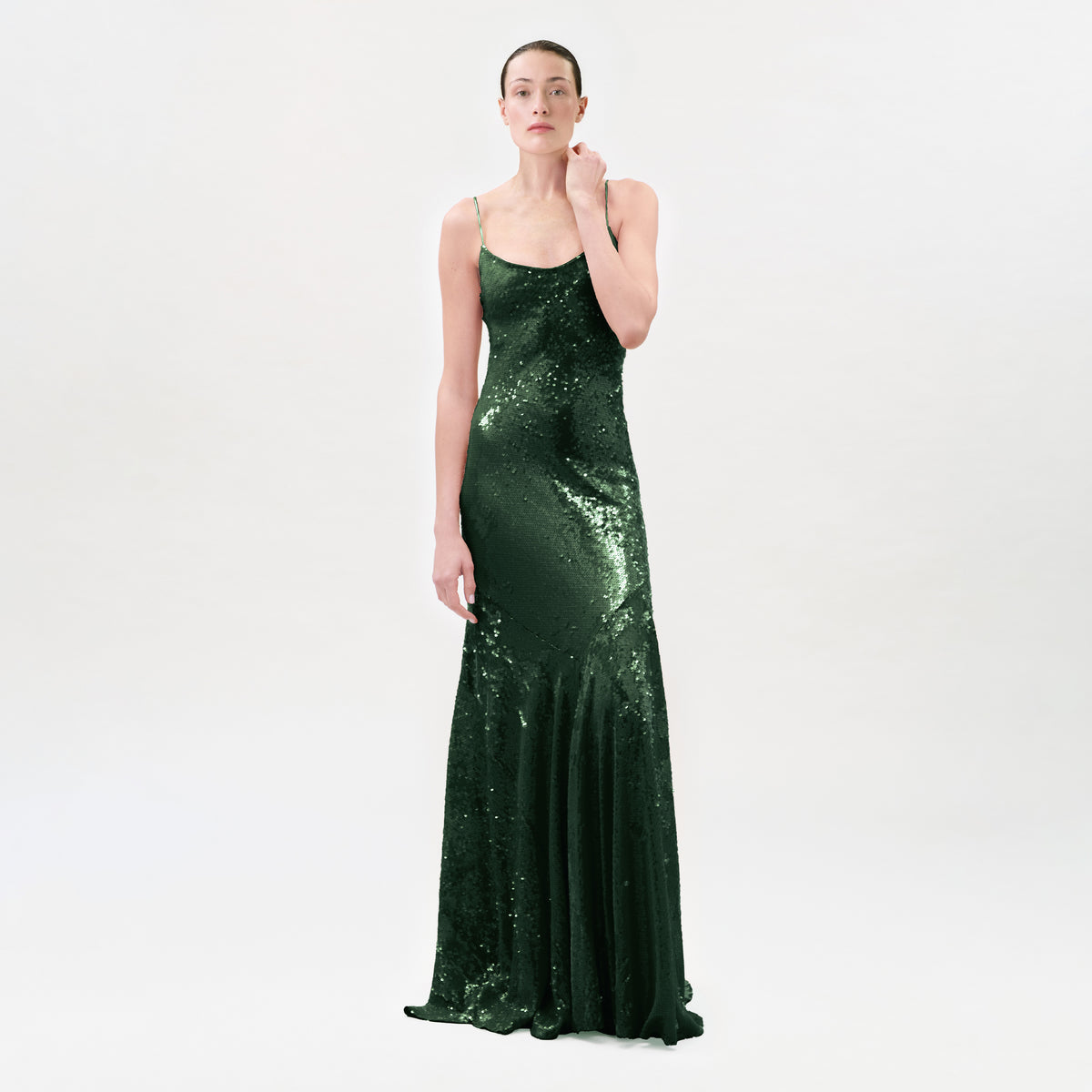 A woman modeling a floor-length, dark green sequined gown with thin spaghetti straps and a fitted silhouette, photographed against a seamless light gray studio background.