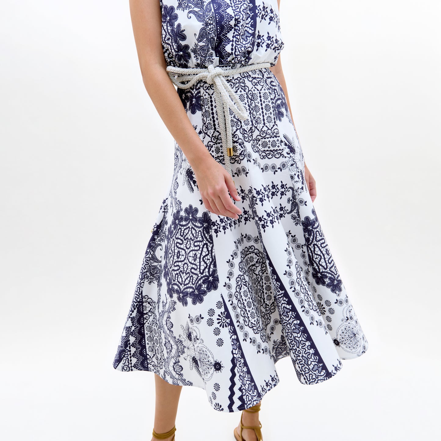 A woman wears the Idona Dress, a sleeveless white midi day dress with blue bandana-style prints and a rope belt at the waist, paired with brown sandals. The image shows her from shoulders to mid-calf.