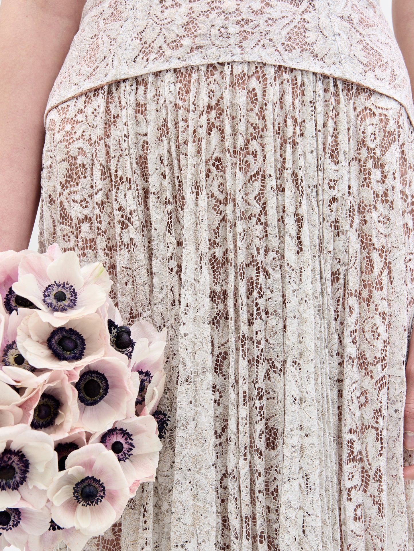 Wearing the Ilana Dress, a woman in a long lace bridal gown holds a bouquet of white and purple flowers against a plain background.