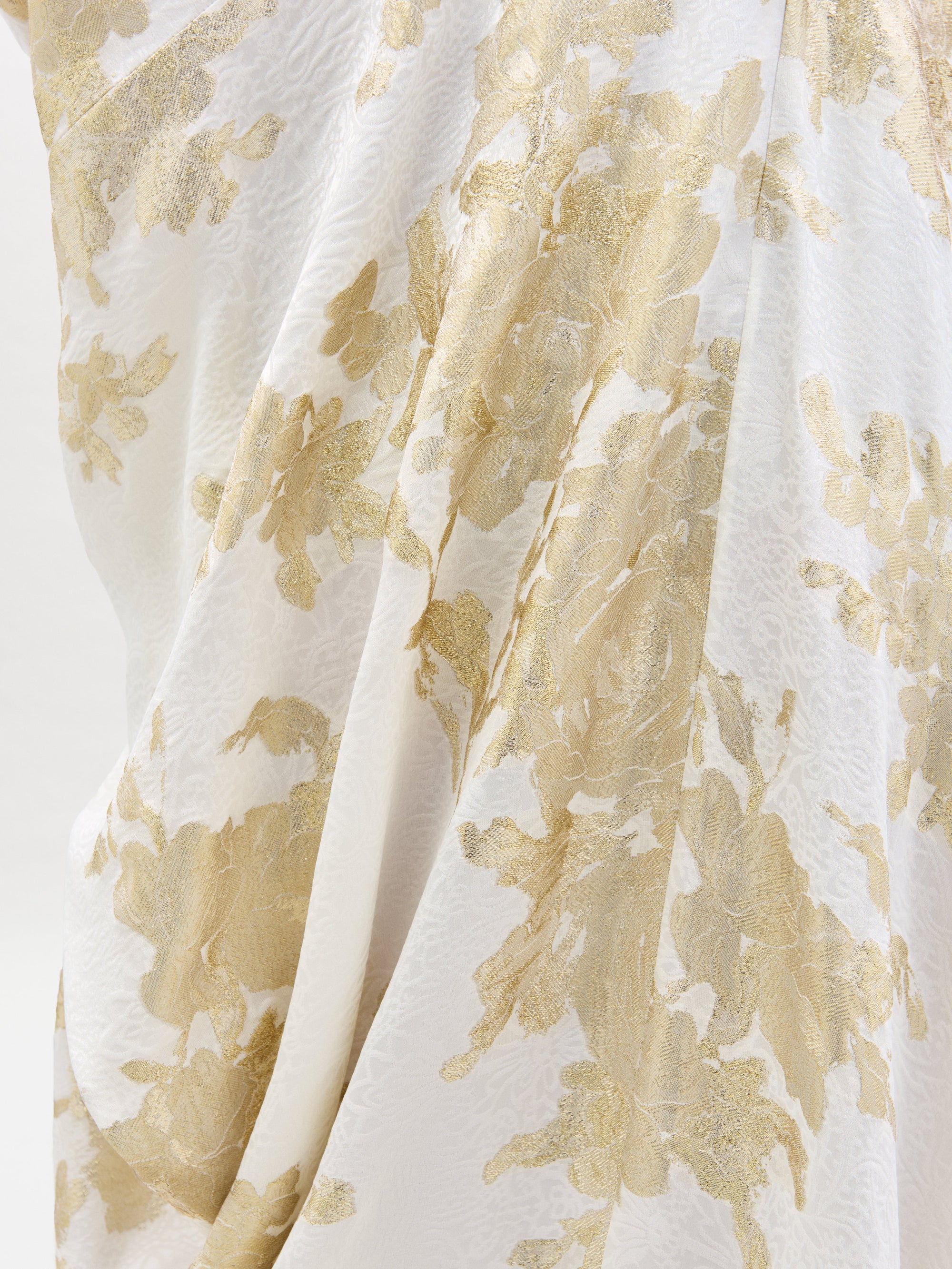 A woman dons the elegant Inu Dress, a flowing white and gold attire with a floral pattern, standing against a plain white background.