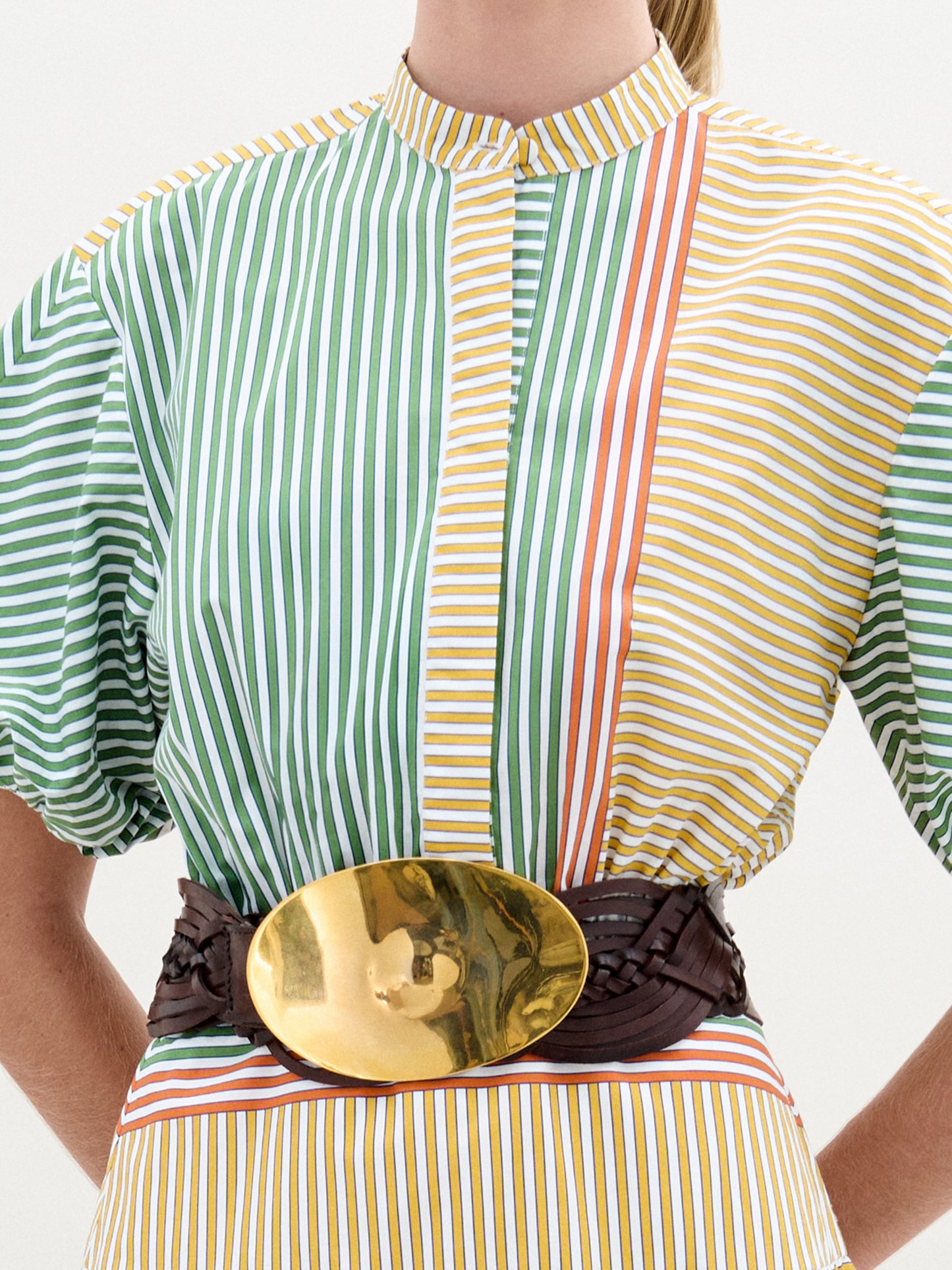 A woman stands against a plain background wearing the Jil Dress, a green and yellow striped midi with puffed sleeves, an A-line skirt, wide belt with large buckle, and gold sandals.