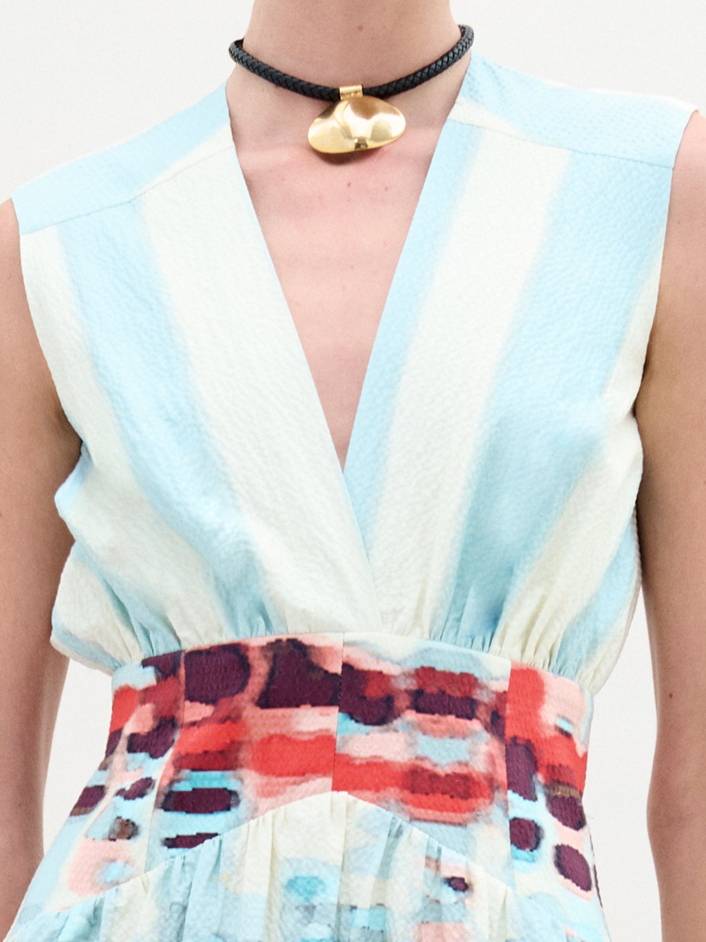 A woman poses against a plain backdrop in the Jocelyn Dress, a sleeveless V-neck midi made from fine Italian fabric with blue stripes and red patterns. She accents the look with gold sandals, a choker, and carries a beige handbag.