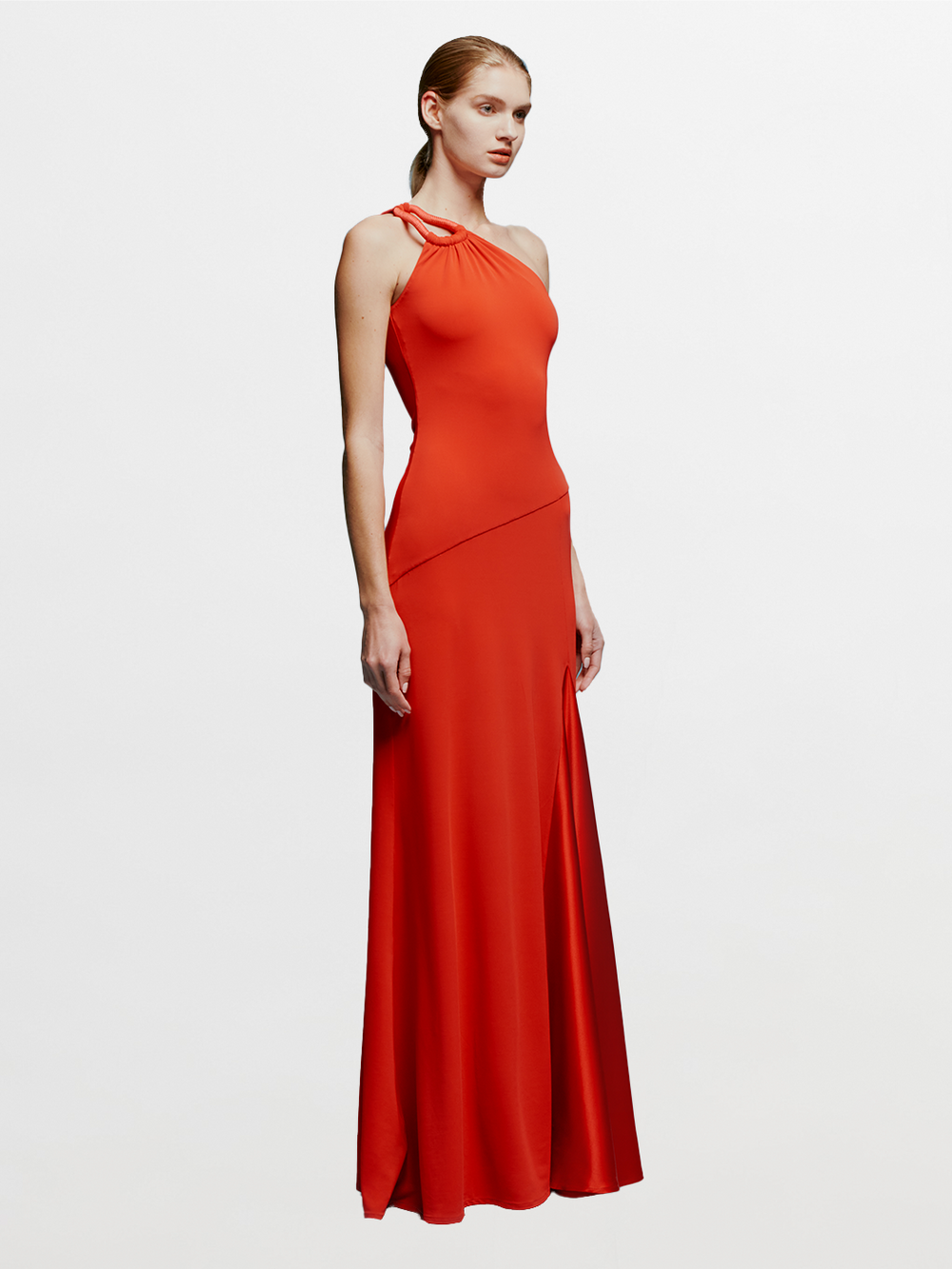 A woman in a Kanya Dress Rouge, featuring a red, one-shoulder design and floor-length cut, stands against a plain blue background. Her hair is pulled back as she calmly stands with her arms by her sides, appearing to wait for an important date such as August 19th or September 2nd.