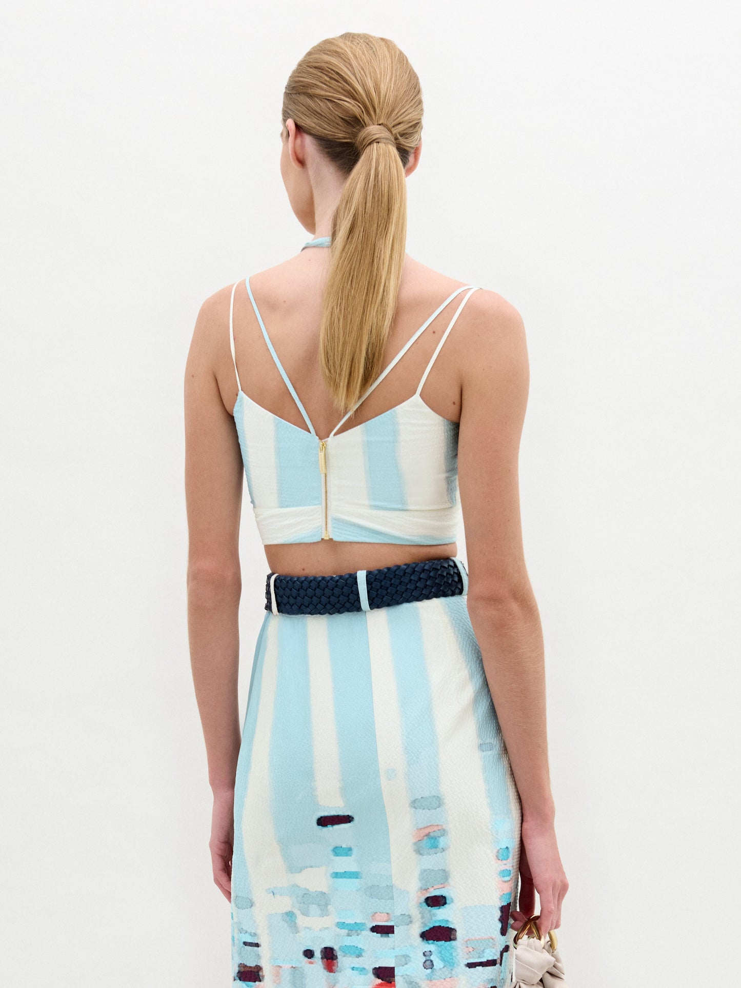 A woman stands against a plain background wearing the Kim Top— a sleeveless crop top with striking 3D floral detailing in red and blue—paired with a light blue patterned skirt.