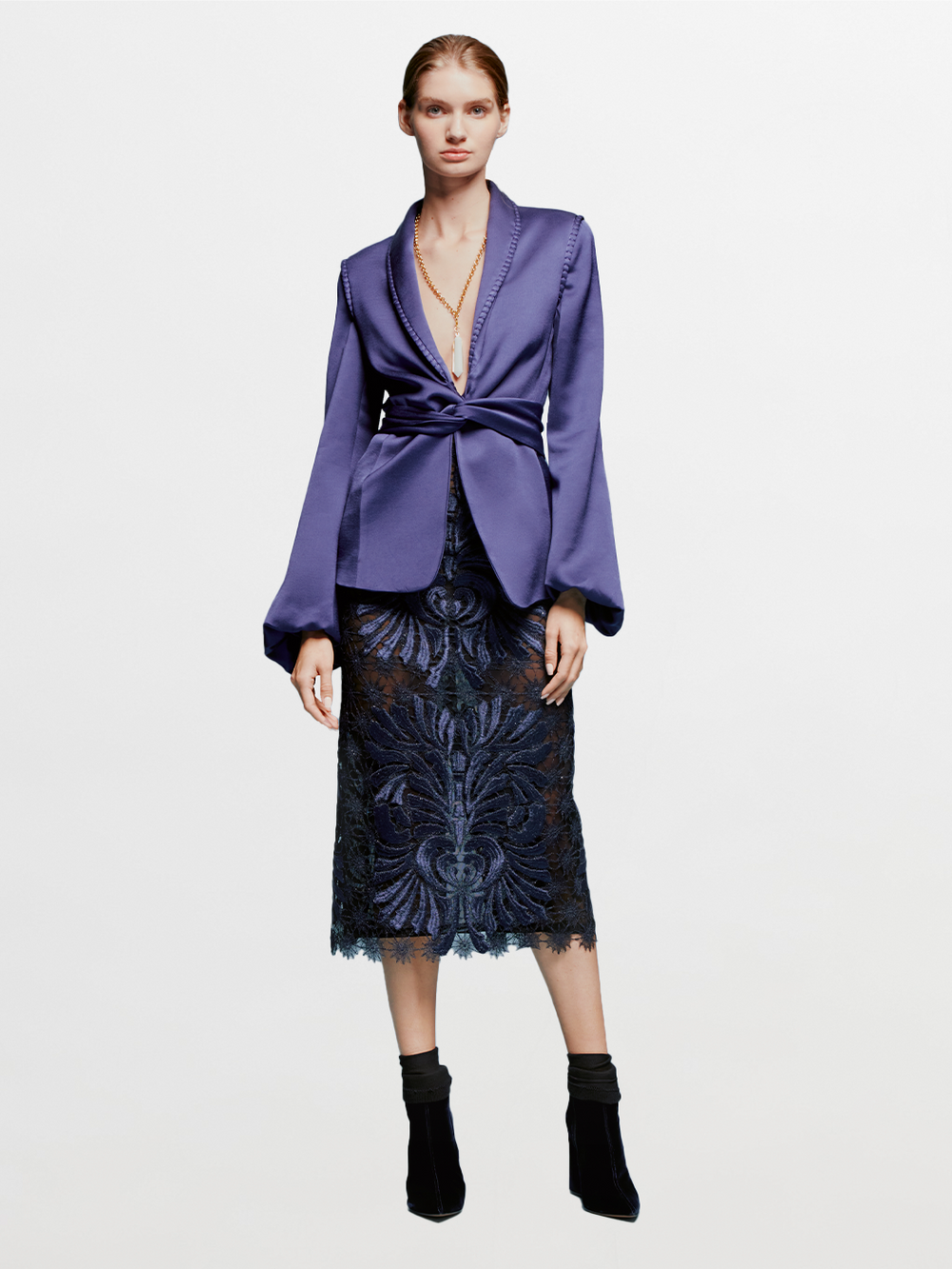 A person stands against a blue background wearing a dark purple jacket over the Kin Skirt Navy, with black heeled boots, ready for the product availability by Ship date August 2024.