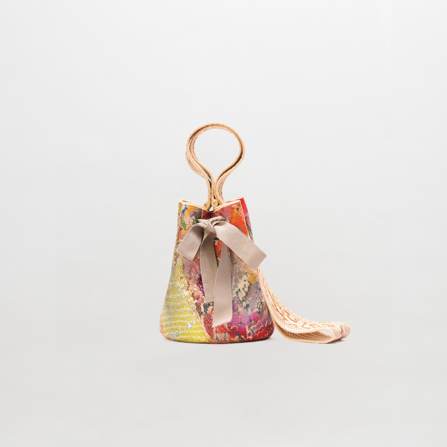 The Mochila Laaput Small is a multicolored patterned bucket bag with a circular handle, beige ribbon detail at the front, and a long hand-woven strap, shown against a plain light background.