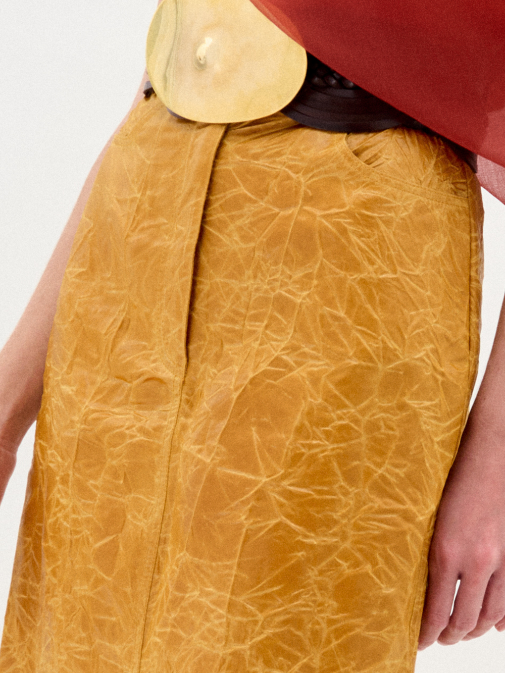 A woman models the Layan Skirt—a mustard yellow structured skirt with a large circular belt—styled with a red blouse and black heeled boots against a plain white background.