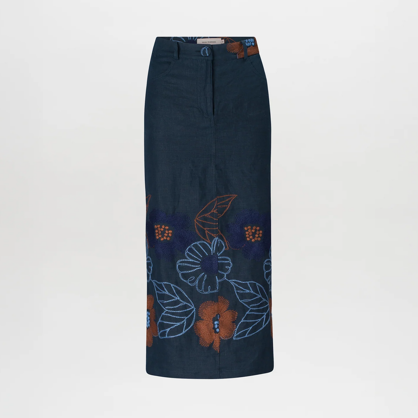 Against a plain background, a woman stands wearing a brown checkered jacket, the Layan Skirt Navy Ochre Flower Embroidery, a brown belt, and gray heeled boots. With her hair tied back neatly, she exudes grace as if ready to board an elegant ship setting sail on August 19th.