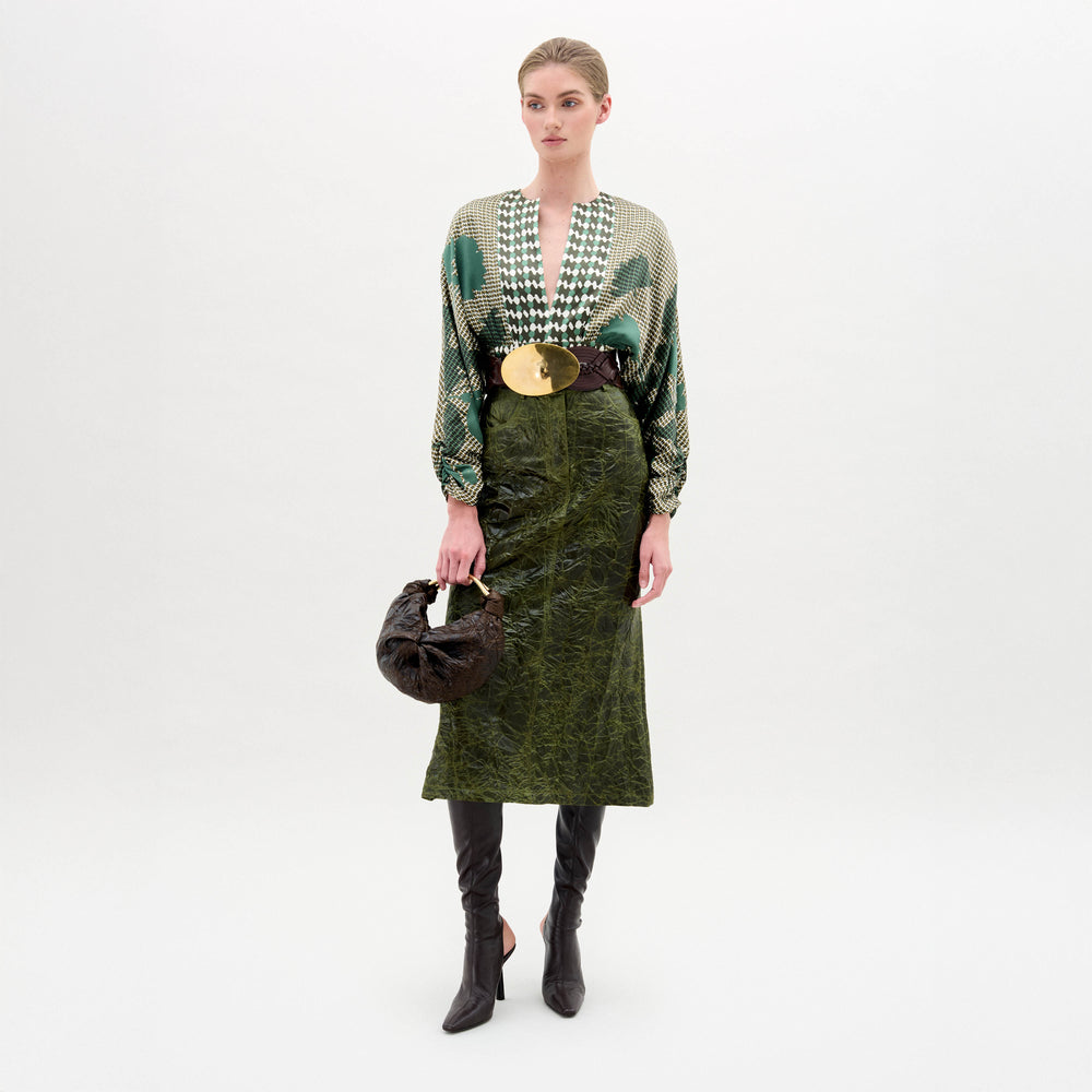 A woman stands against a plain background wearing a patterned green blouse, the Layan Skirt, black boots, and holding a round black bag.