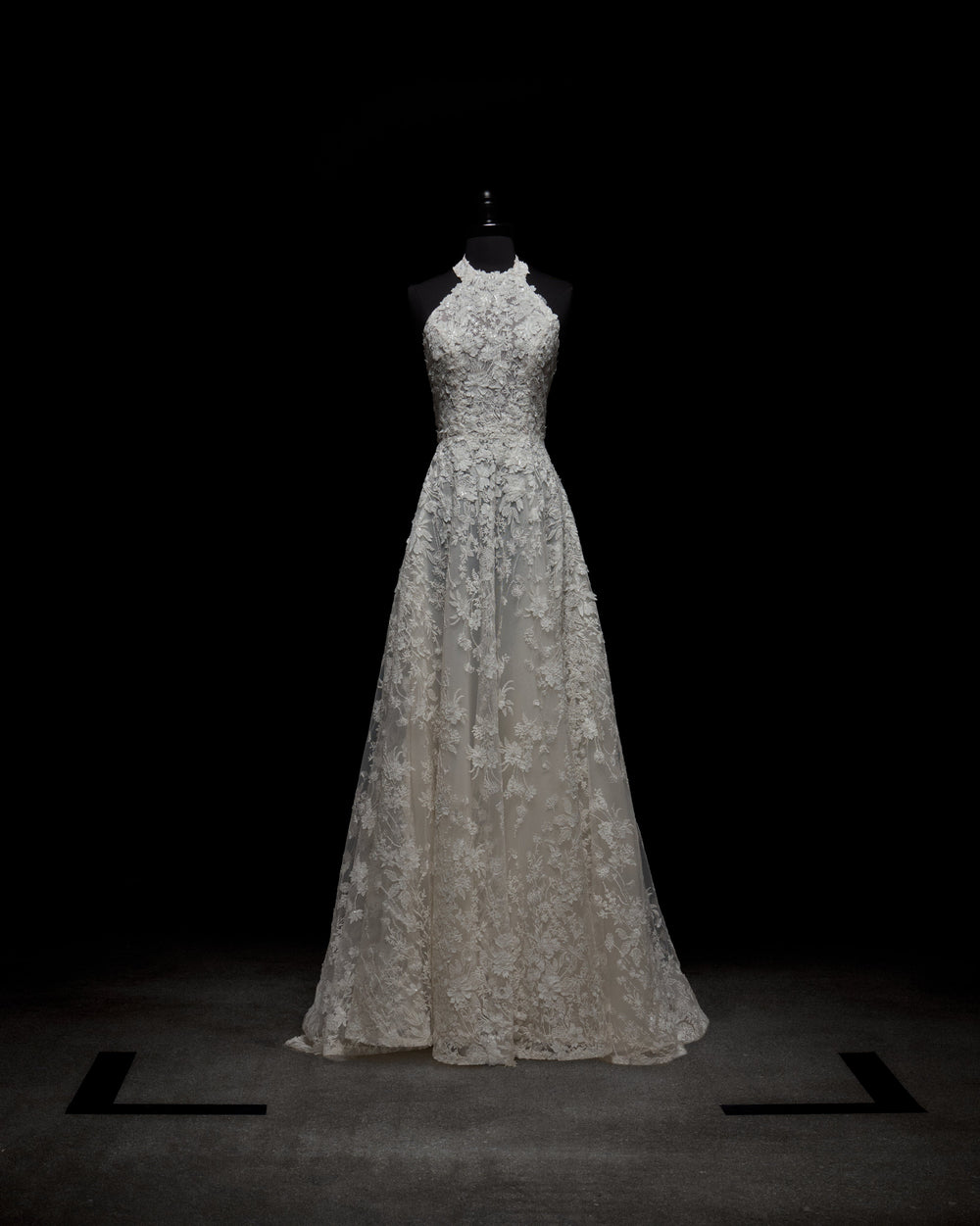 A woman in the sleeveless Leia Dress, a white lace wedding gown, stands against a plain white background with her hands behind her back.