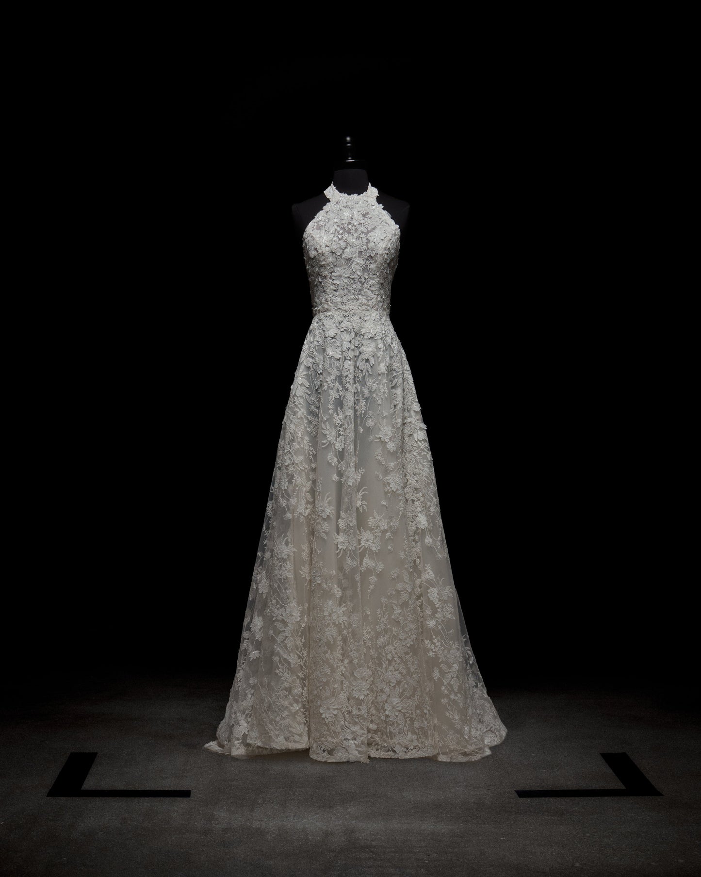 A woman in the sleeveless Leia Dress, a white lace wedding gown, stands against a plain white background with her hands behind her back.