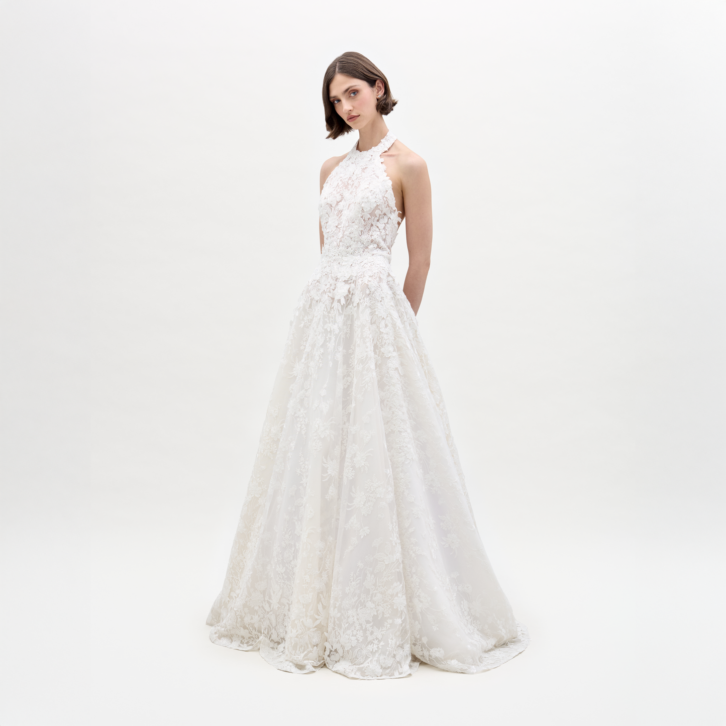 A woman in the sleeveless Leia Dress, a white lace wedding gown, stands against a plain white background with her hands behind her back.