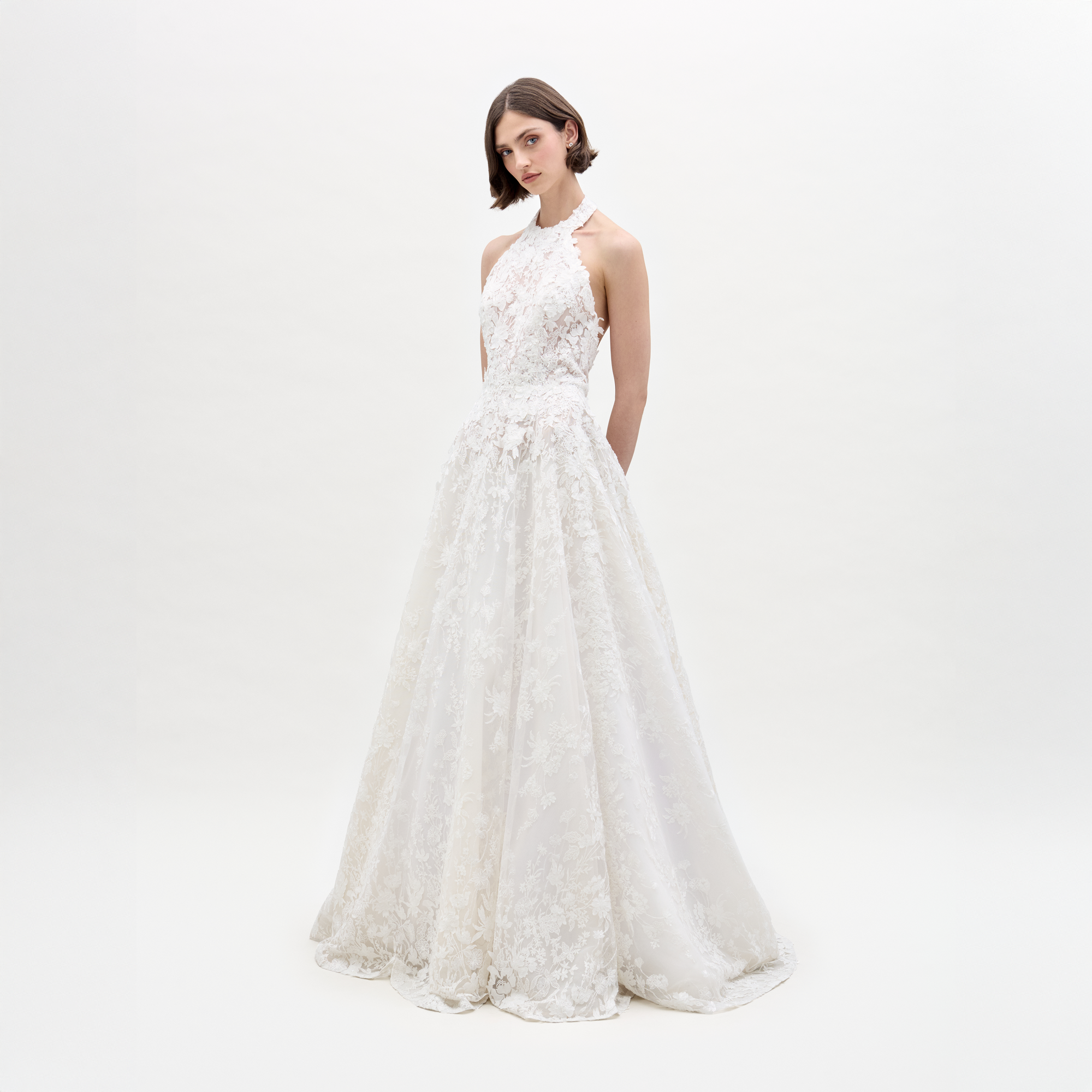 A woman in the sleeveless Leia Dress, a white lace wedding gown, stands against a plain white background with her hands behind her back.