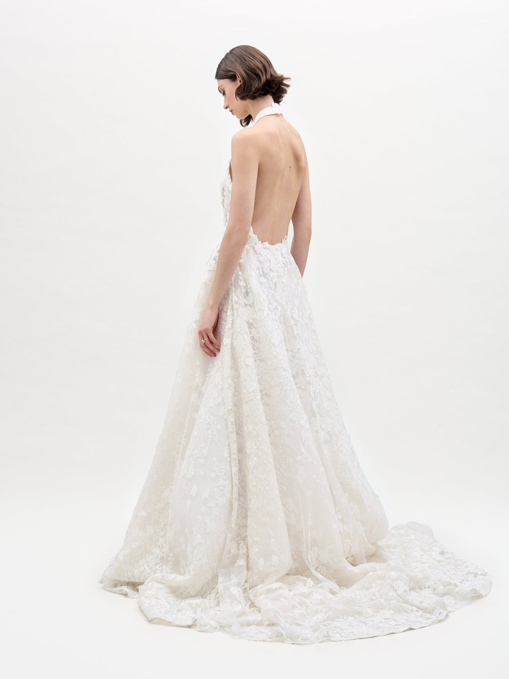 A woman in the sleeveless Leia Dress, a white lace wedding gown, stands against a plain white background with her hands behind her back.