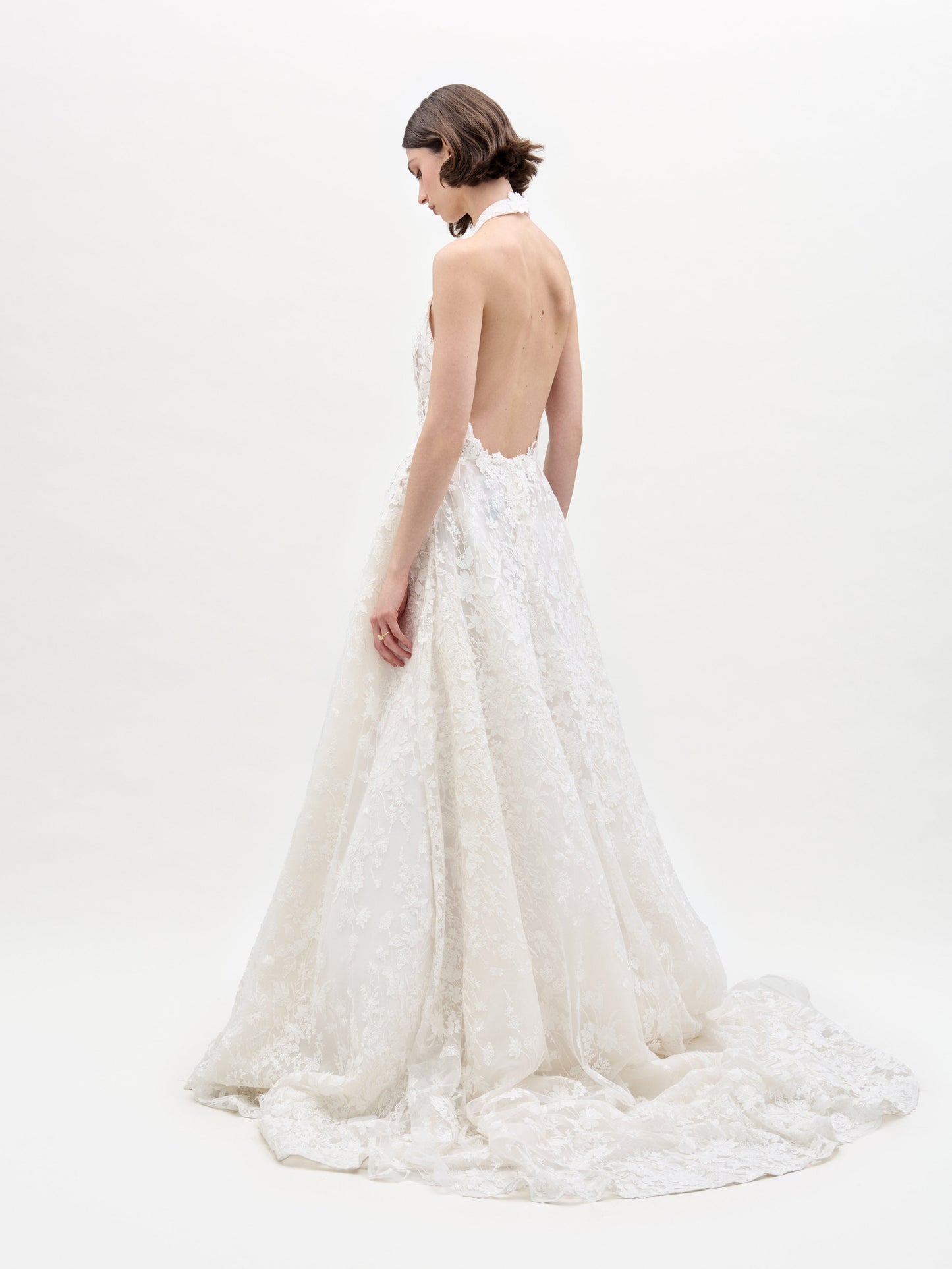 A woman in the sleeveless Leia Dress, a white lace wedding gown, stands against a plain white background with her hands behind her back.