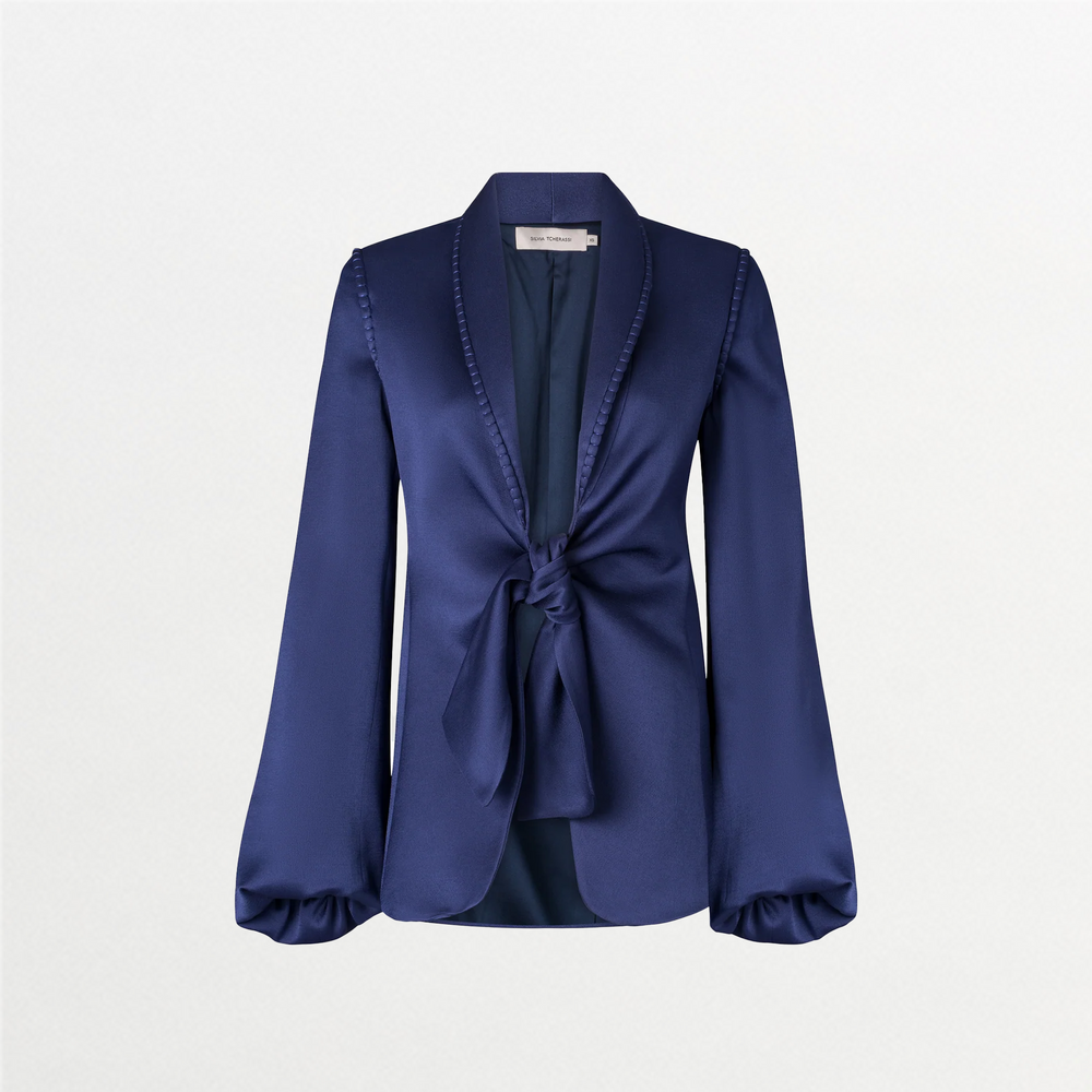 A person in a chic Leo Jacket Navy and a patterned skirt stands against a light blue background, embodying the stylish essence of September 2nd. They are wearing a long necklace with a white pendant.