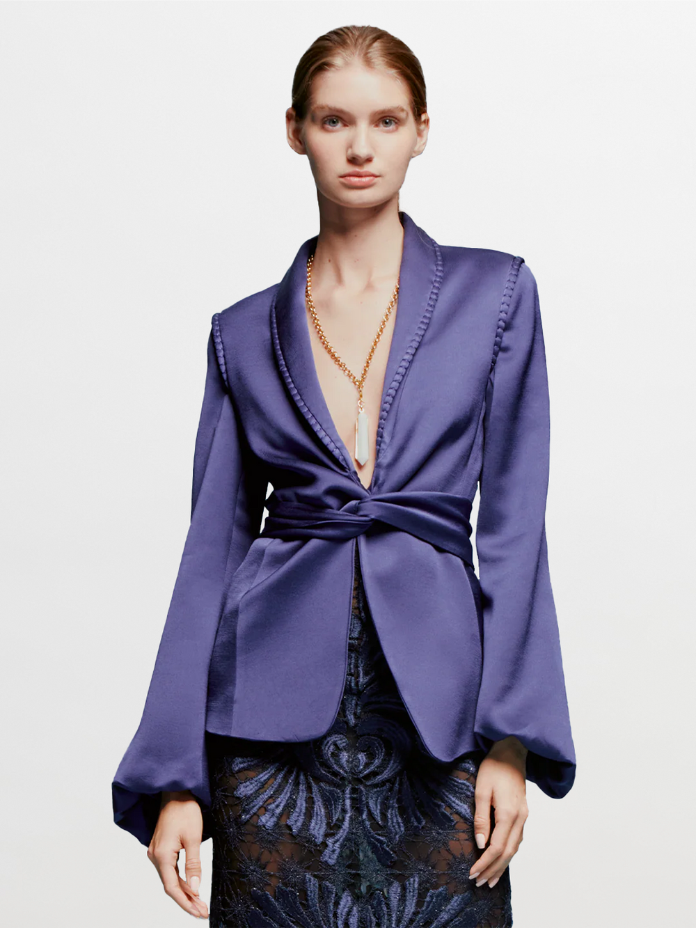A person in a chic Leo Jacket Navy and a patterned skirt stands against a light blue background, embodying the stylish essence of September 2nd. They are wearing a long necklace with a white pendant.