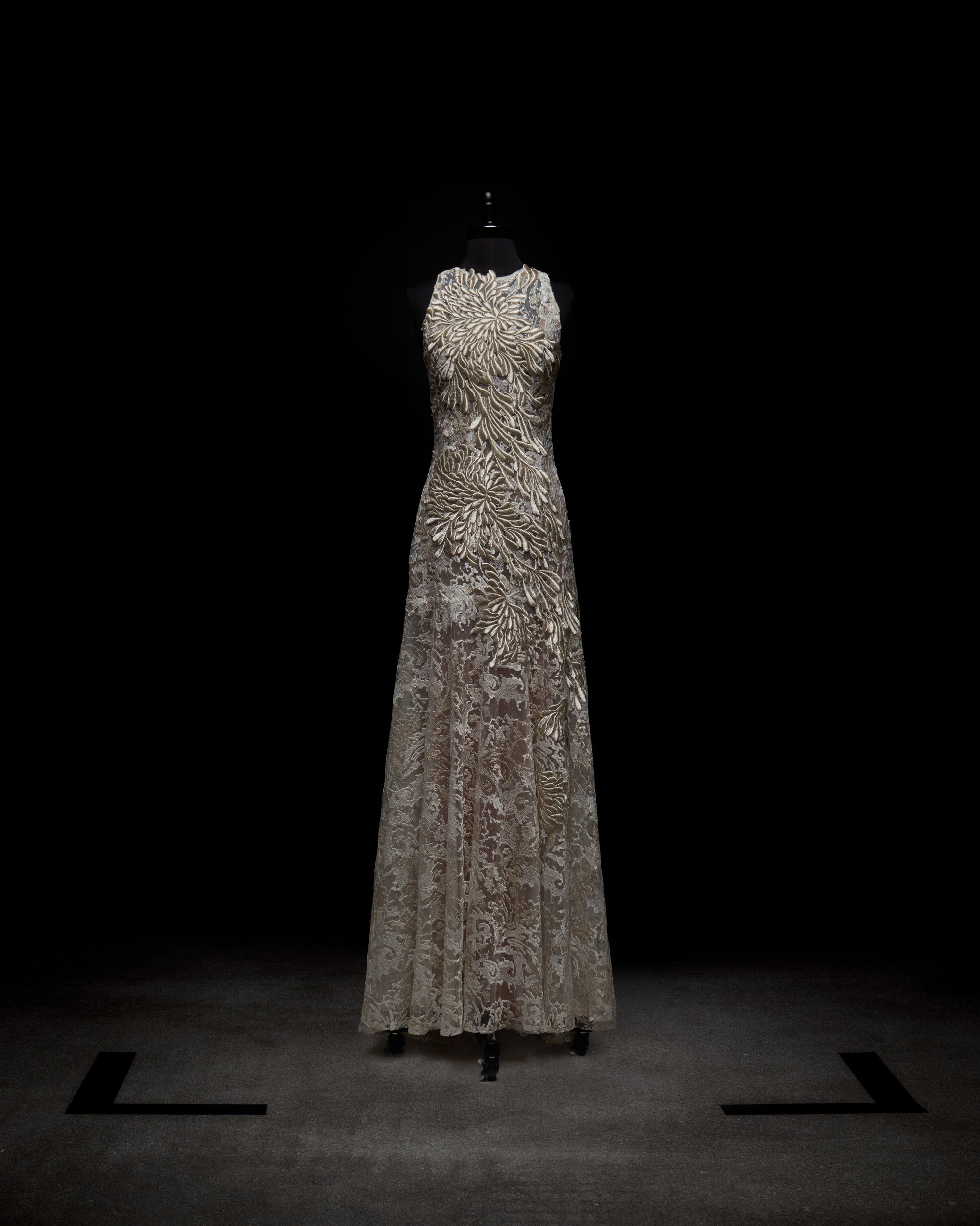 A woman models the Levi Dress, a sleeveless, floor-length maxi featuring floral embroidery and delicate appliqué, standing against a plain white background.