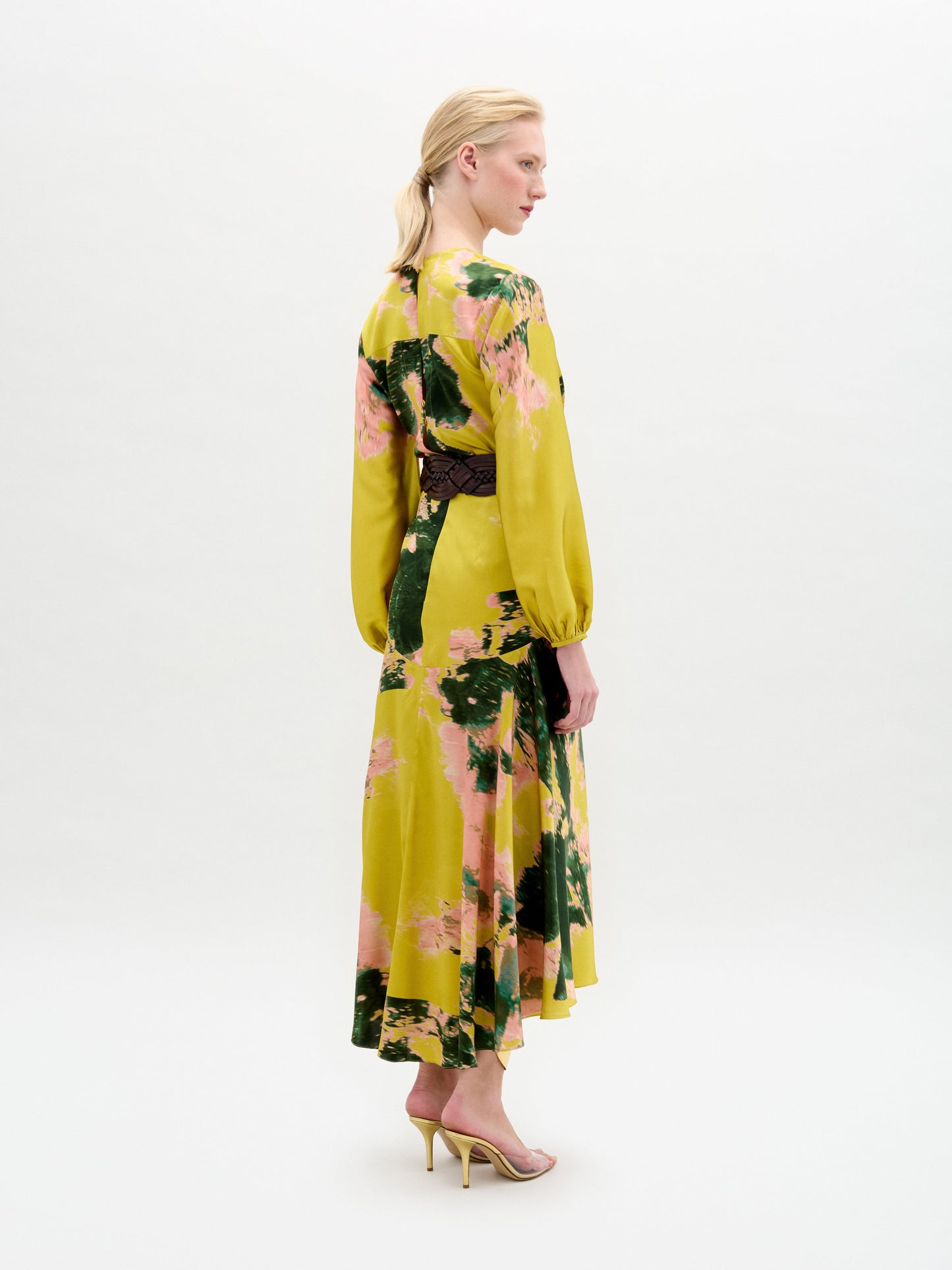 A person in a Lima Dress with a floral yellow and green pattern, a wide belt, holding a beige handbag, and wearing strappy high heels.