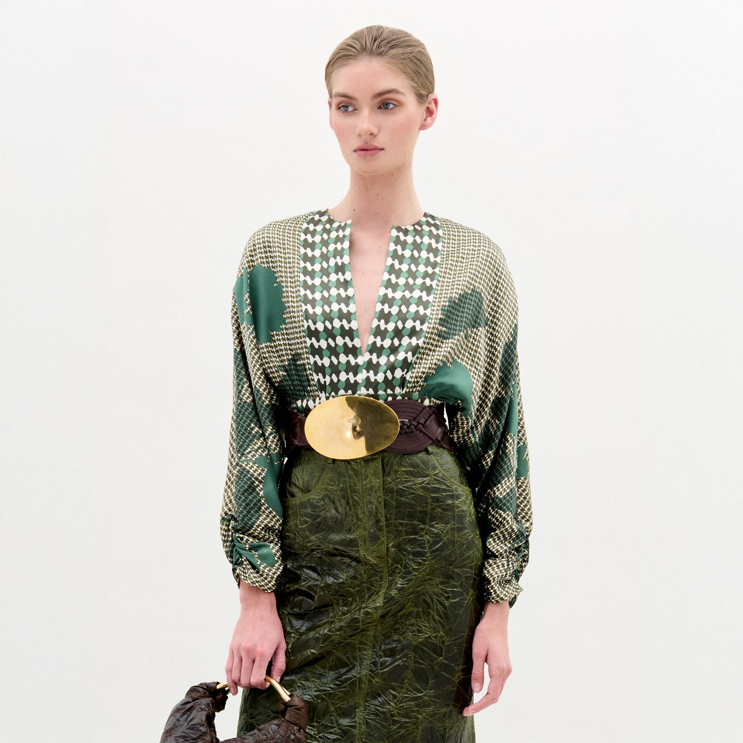A woman stands against a white background wearing the Lizzy Blouse, a green silk patterned top, paired with a dark green textured skirt, wide belt with large gold buckle, and holding a brown bag.