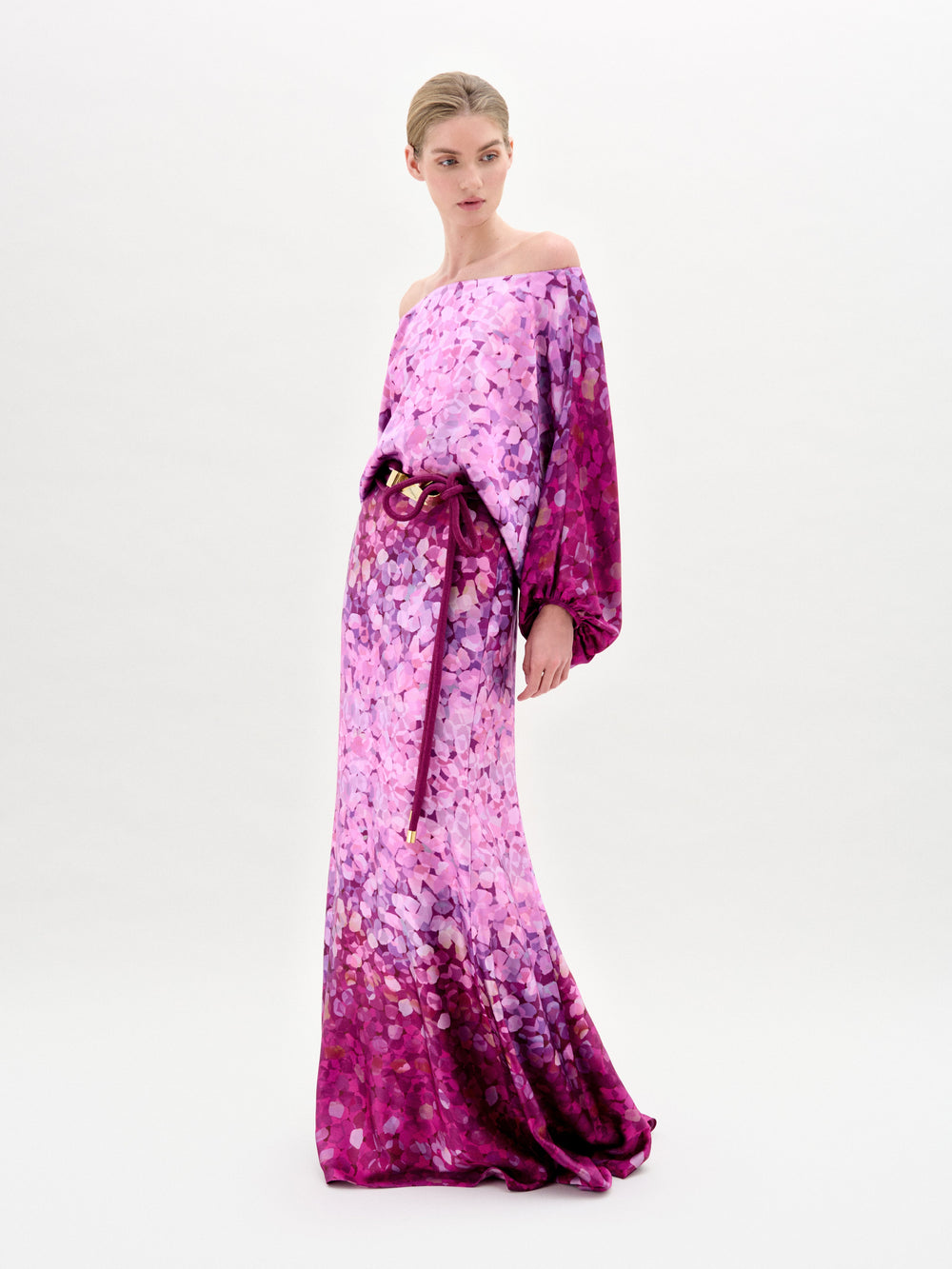 Woman models the Lorella Dress—an off-the-shoulder maxi with purple and pink florals, crafted from luxurious Italian fabric and finished with a tied belt—against a plain white background.