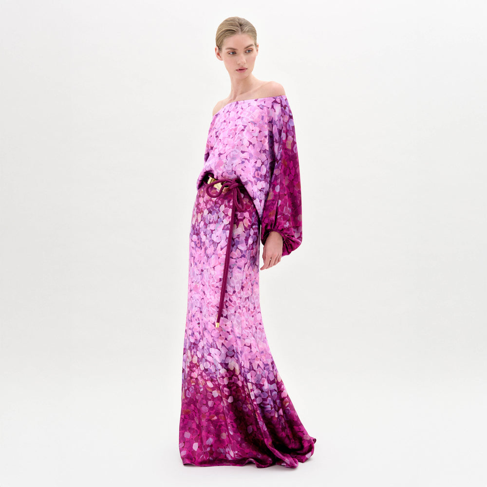A person wearing the Lorella Dress, an off-the-shoulder maxi dress crafted from luxurious Italian fabric and adorned with a purple, pink, and magenta floral pattern, stands against a plain background.