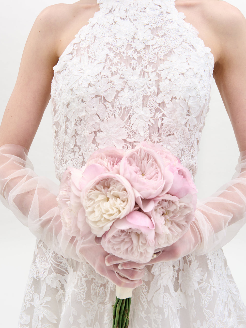 A woman in the sleeveless Leia Dress, a white lace wedding gown, stands against a plain white background with her hands behind her back.