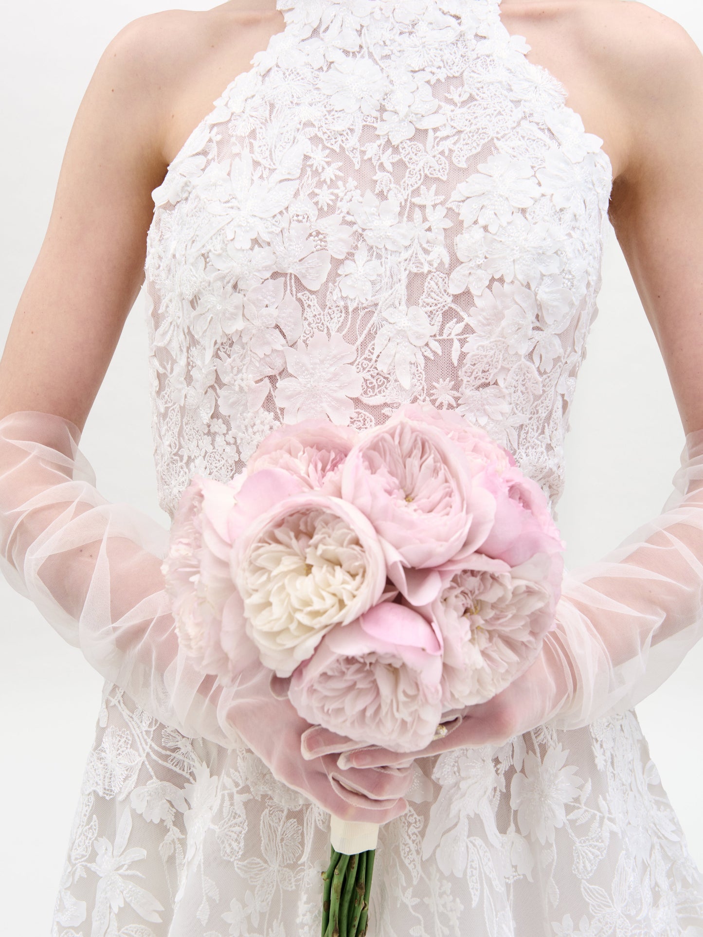 A woman in the sleeveless Leia Dress, a white lace wedding gown, stands against a plain white background with her hands behind her back.