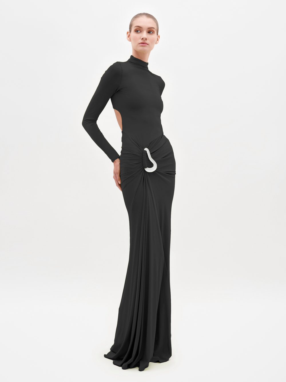 A woman poses against a plain white background wearing the Lena Dress, a long black maxi featuring a high neckline, open back, gathered waist, draped skirt, and white decorative detail at the hip.
