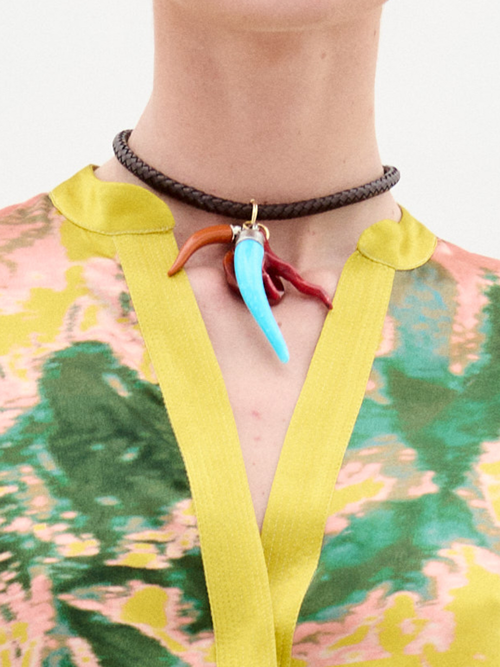 The Leslie Chocker is a brown braided bracelet with gold metallic accents, multicolor horn-shaped charms in red, blue, and brown, and a large red faceted bead displayed on a white background.