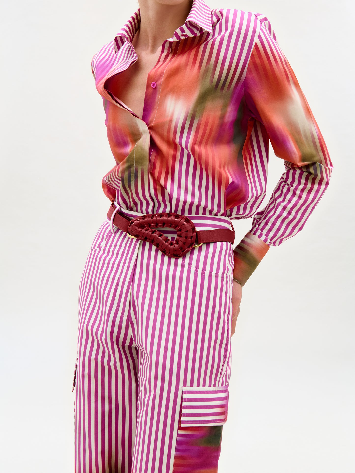 A person wearing the Linette Blouse, a long-sleeve Italian fabric top with a pink and orange blurred pattern, pink and white striped cotton pants, and a braided red belt stands against a plain light background.