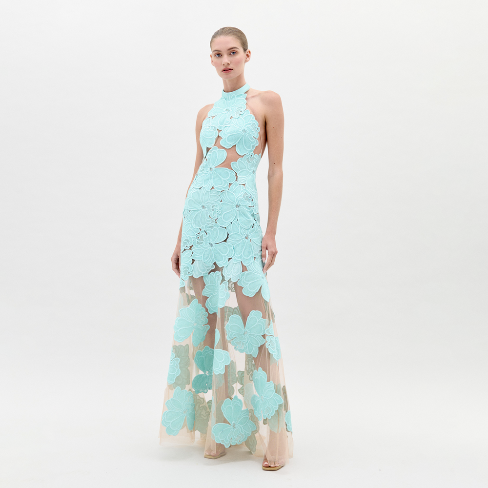 A woman wearing the Mab Dress Turquoise, a light blue floral-patterned halter neck dress with a sheer skirt, stands against a plain background, eagerly awaiting the pre-order ship-by date of February 1st.