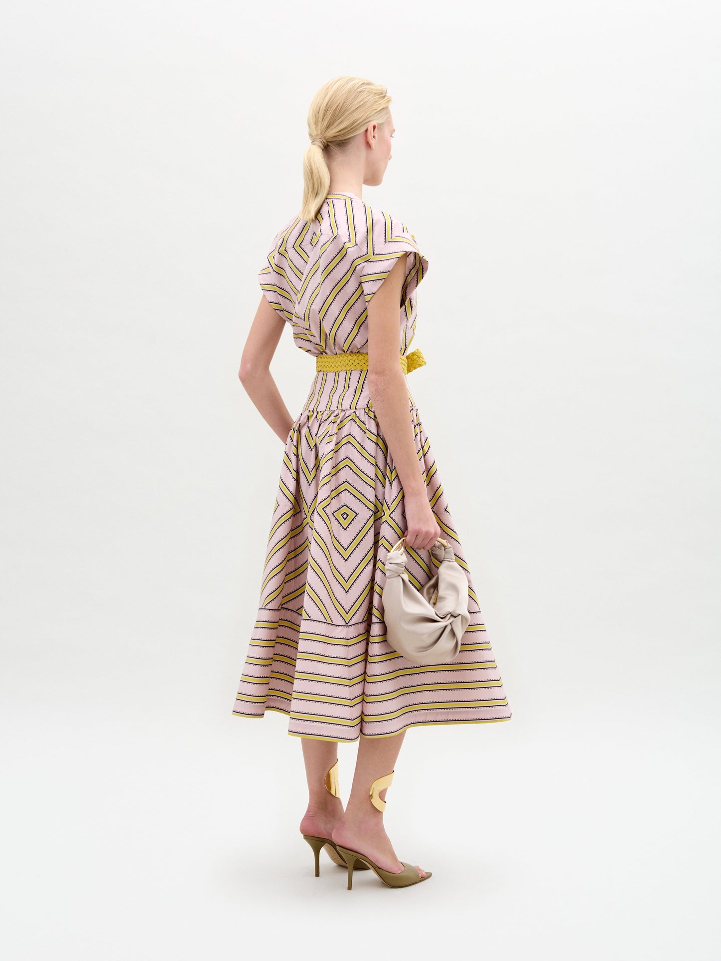 A woman models the Macarena Dress—a light pink and yellow geometric midi made from Italian fabric—styled with a woven leather belt and high-heeled sandals, set against a plain white background.