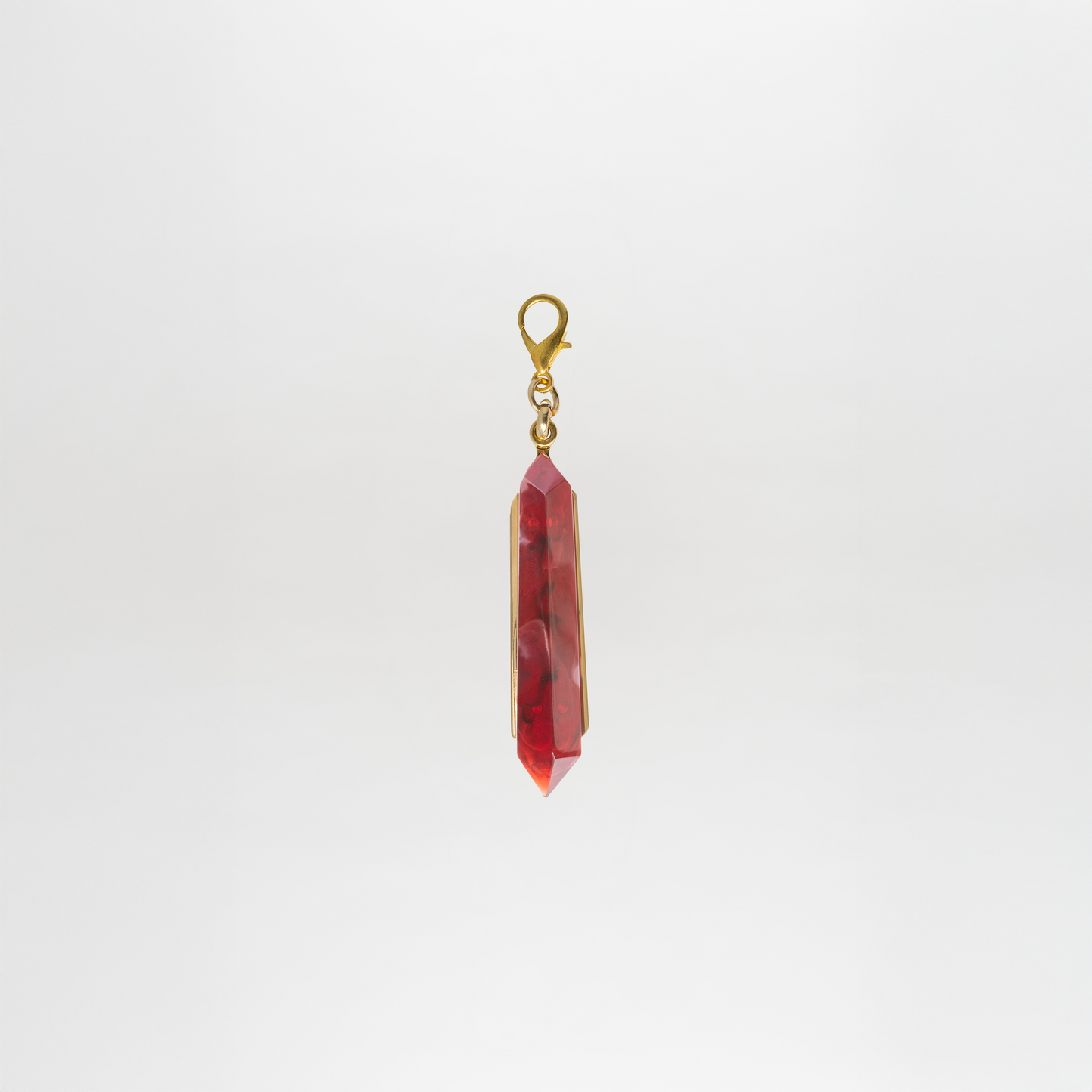 The Mariela Charm features a gold plated brass clasp holding a red faceted crystal pendant, suspended vertically against a plain white background.