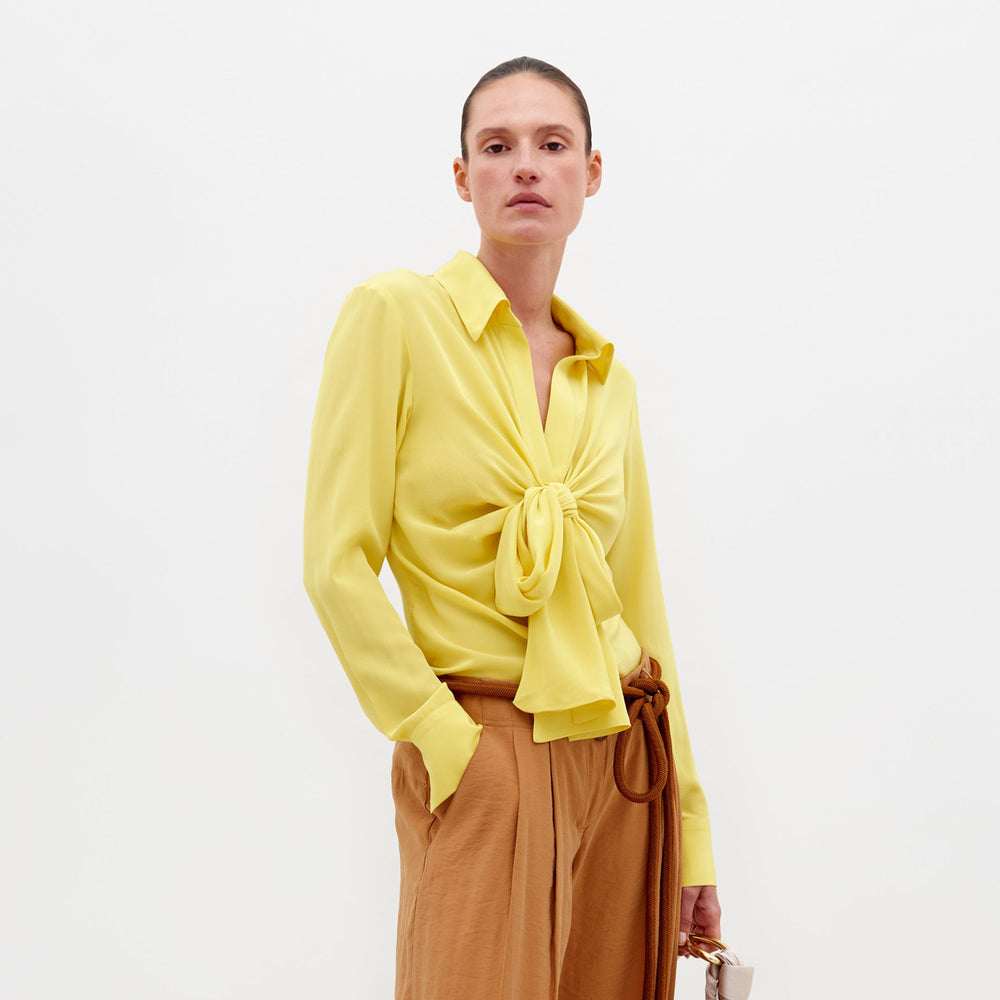 An individual stands against a plain backdrop wearing the Mercury Blouse Yellow, featuring long sleeves and a knotted front, paired with light brown belted trousers. They hold a small beige bag in their left hand. Pre-order now to ensure delivery by February 15th, 2025.