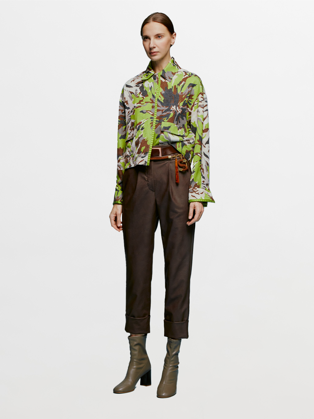 A person is standing against a blue background, wearing a green and brown patterned shirt, dark pants referred to as the "Moad Pant Brown," and gray heeled boots. The shirt is partially tucked into the pants, creating a stylish look perfect for August 19th.