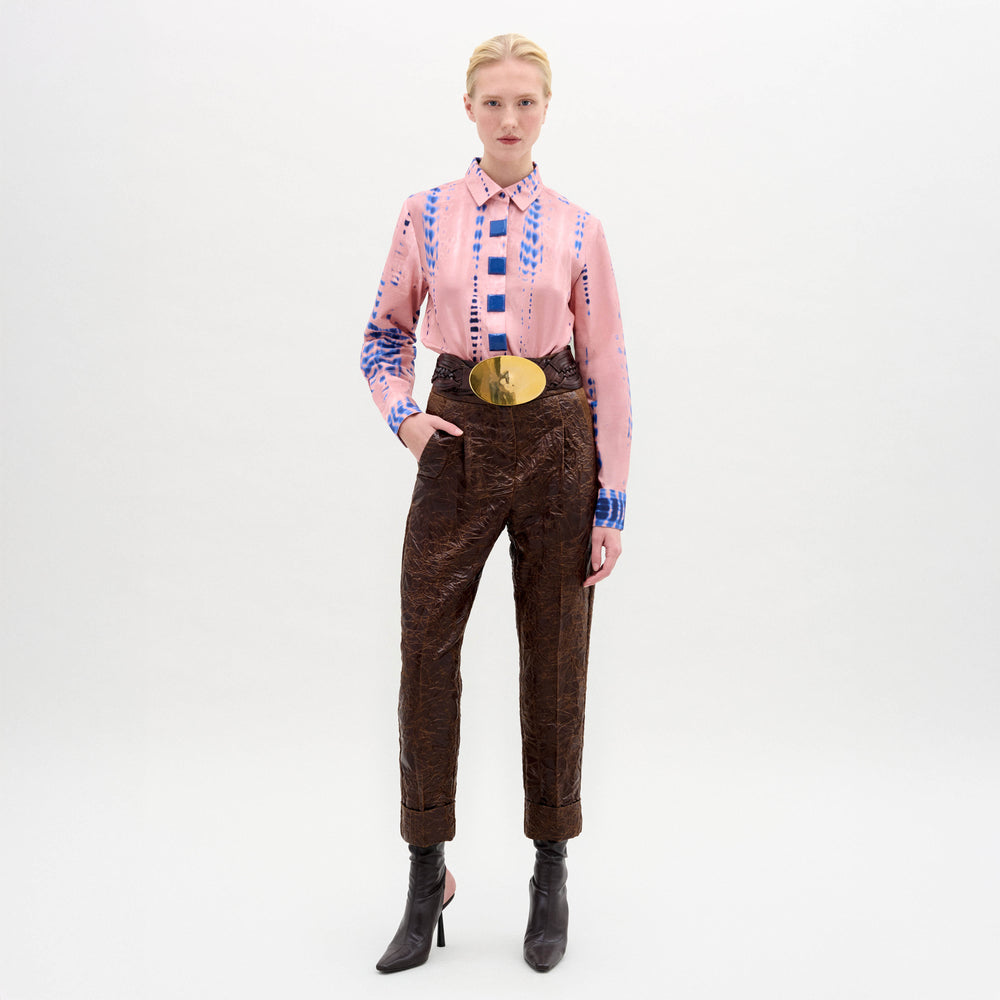 Against a plain backdrop, someone wears the Moad Pant—high-waisted and cropped in Italian fabric with a straight fitted leg—paired with a pink patterned shirt, large gold belt, and black heeled boots.