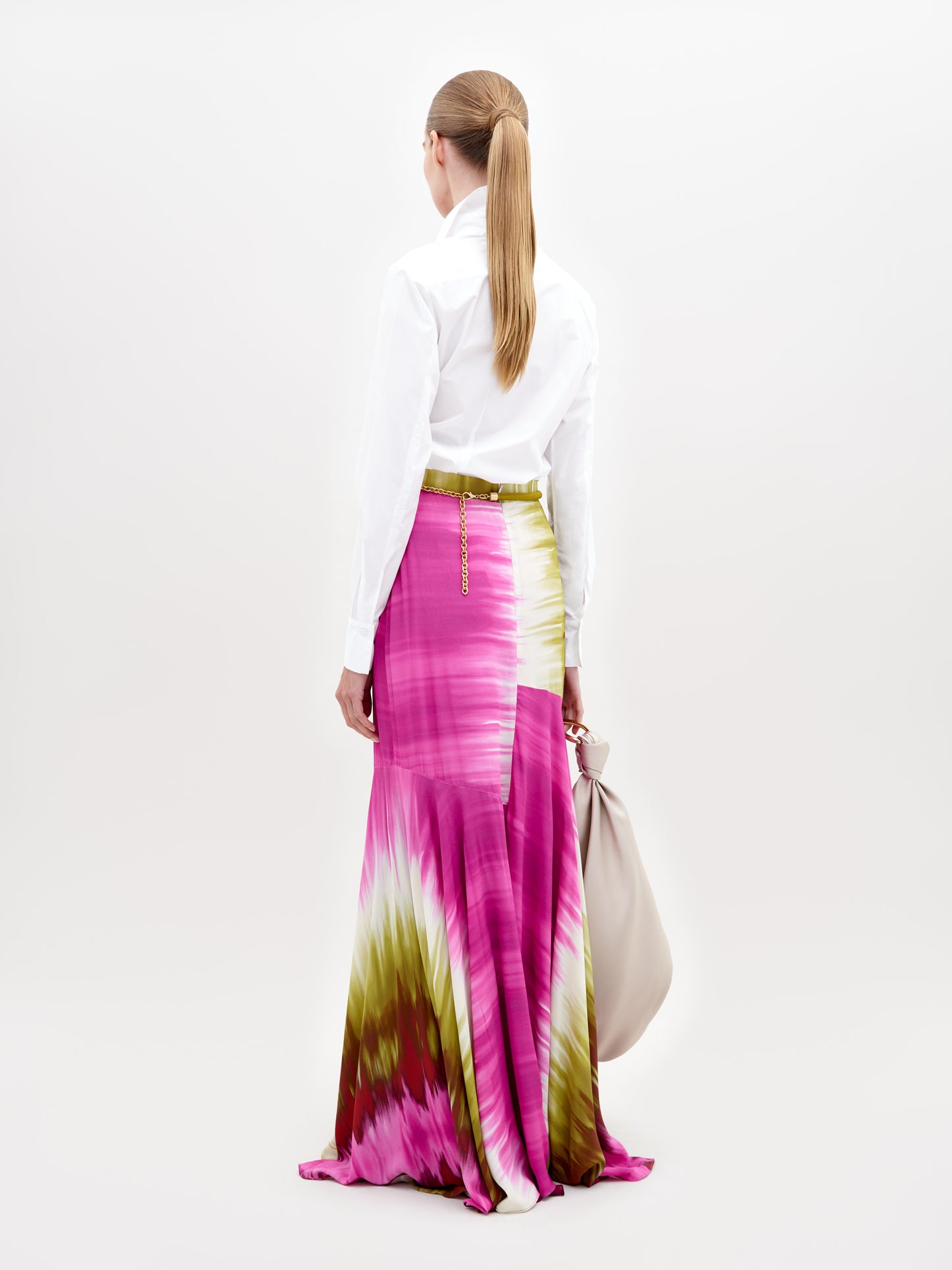 A woman with a ponytail stands facing away, wearing a white long-sleeve shirt and the Shane Skirt—a high-waisted, pink, yellow, and white tie-dye maxi skirt crafted from Italian textile—holding a large light gray bag against a plain white background.