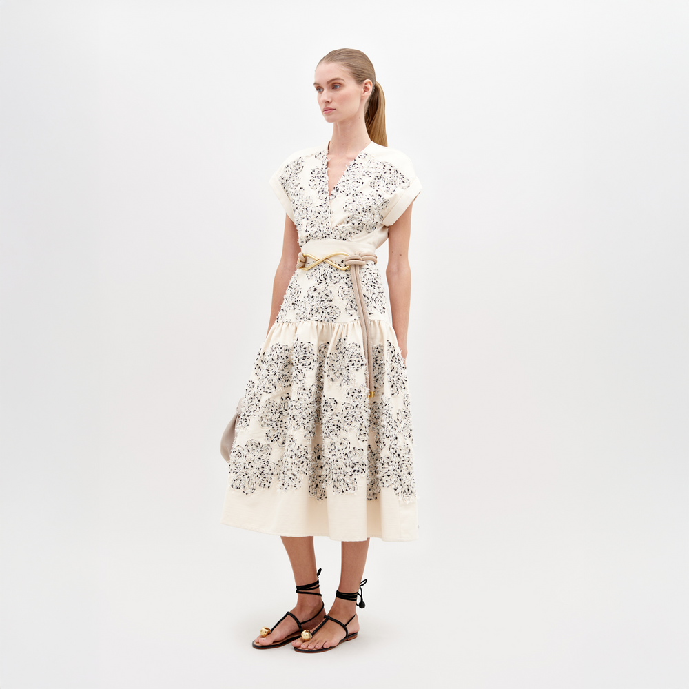 A woman wearing a Metaponto Dress Beige Black Embroidered Flowers with a beige belt and black sandals stands against a plain white background, ready for the fashion pre-order set to ship by date November 15th, 2024.