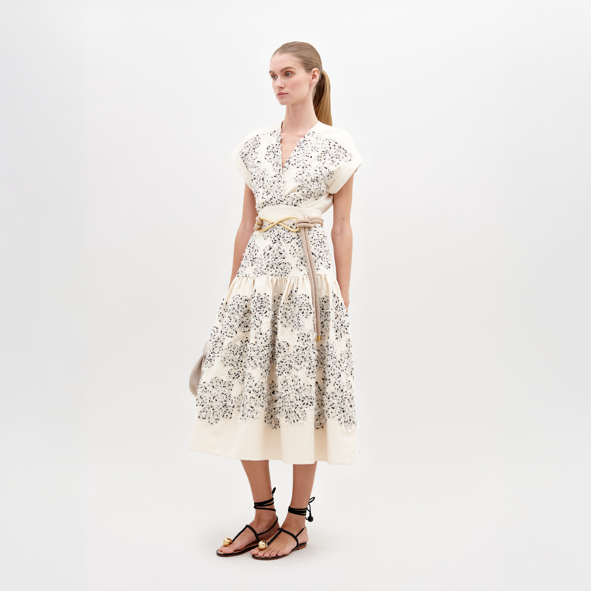 A woman wearing a Metaponto Dress Beige Black Embroidered Flowers with a beige belt and black sandals stands against a plain white background, ready for the fashion pre-order set to ship by date November 15th, 2024.