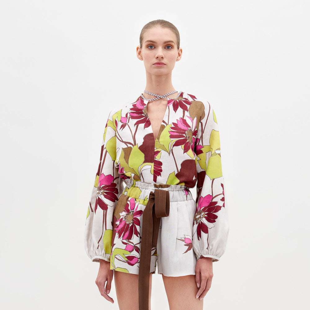 A person stands against a plain background, wearing the Molveno Blouse in Botanical Multifloral, featuring a floral-patterned long-sleeve top with a deep V-neckline and matching high-waisted shorts, cinched at the waist with a brown belt. Pre-order now to ensure it arrives by the February 15th, 2025 ship-by date.