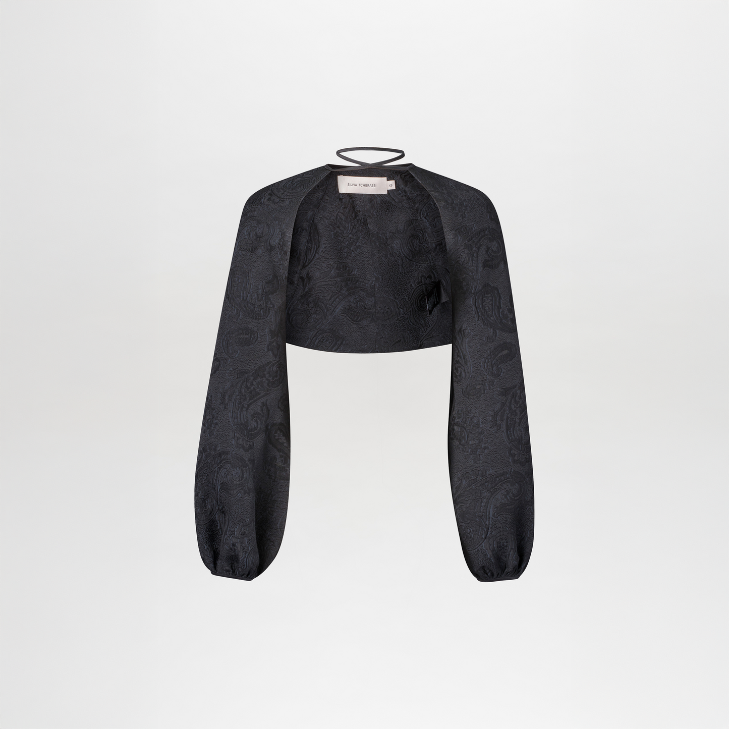 The Nana Cape is a black cropped bolero jacket featuring long, loose sleeves, a subtle jacquard paisley pattern, and a thin neckline strap, shown against a plain white background.